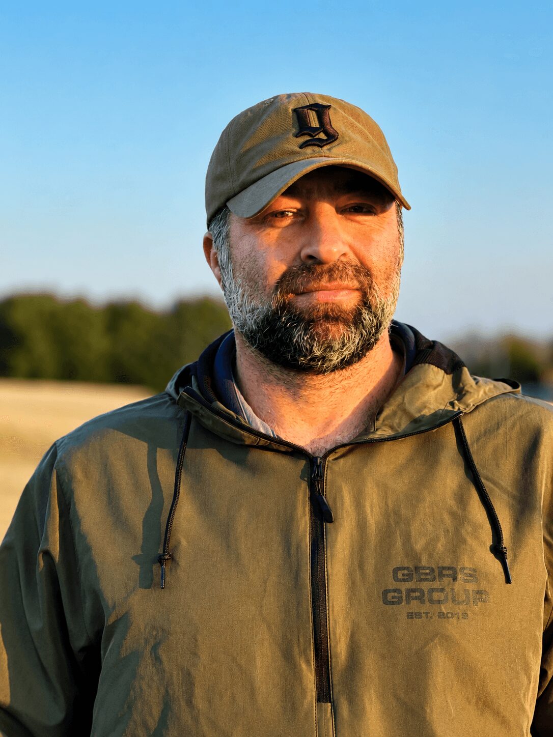 Man with beard wearing a beige cap and green jacket outdoors, smiling slightly, with trees and blue sky in background.