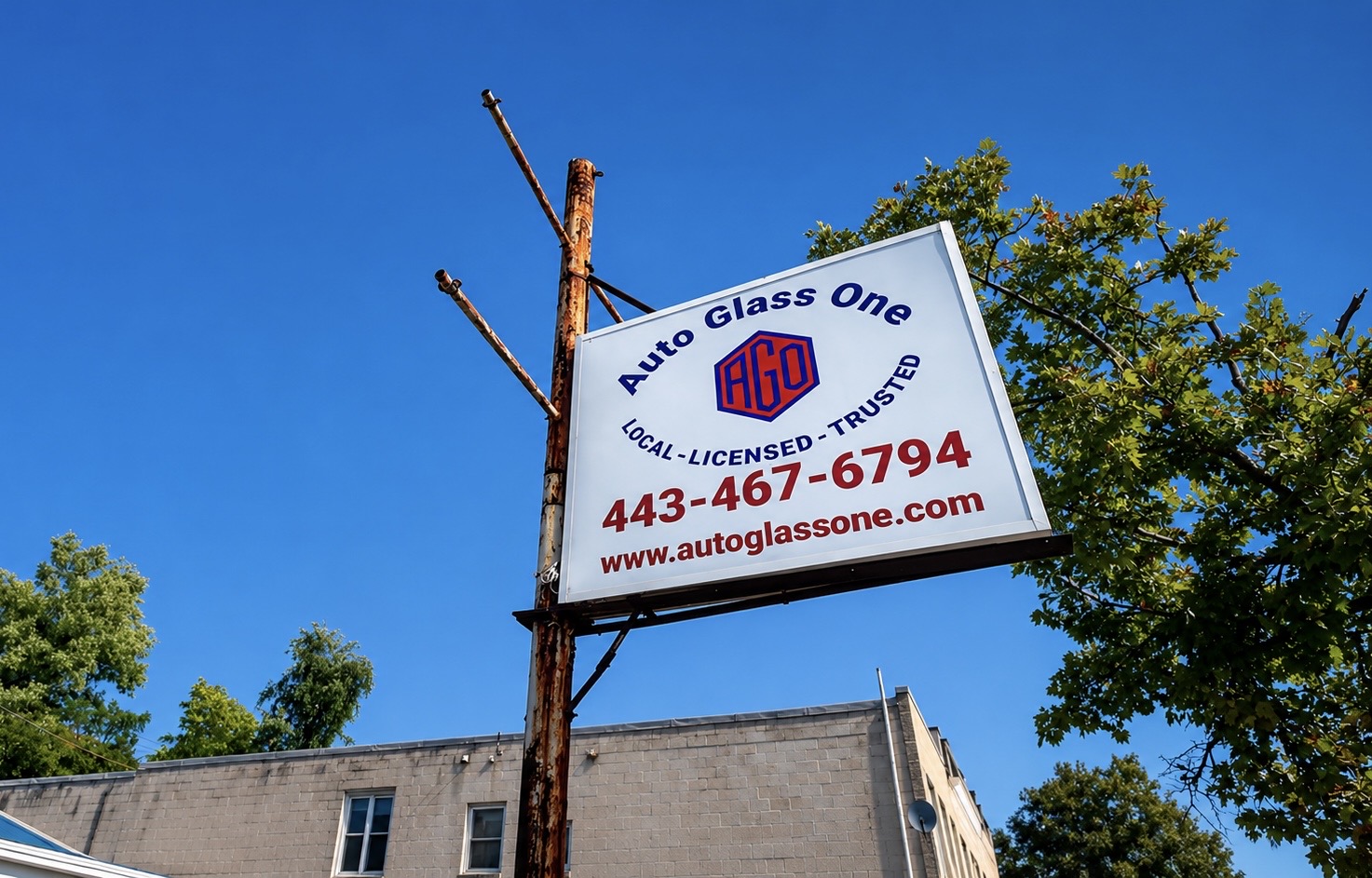 Sign for Auto Glass One with contact number and website, mounted on a wooden pole against a blue sky with trees.