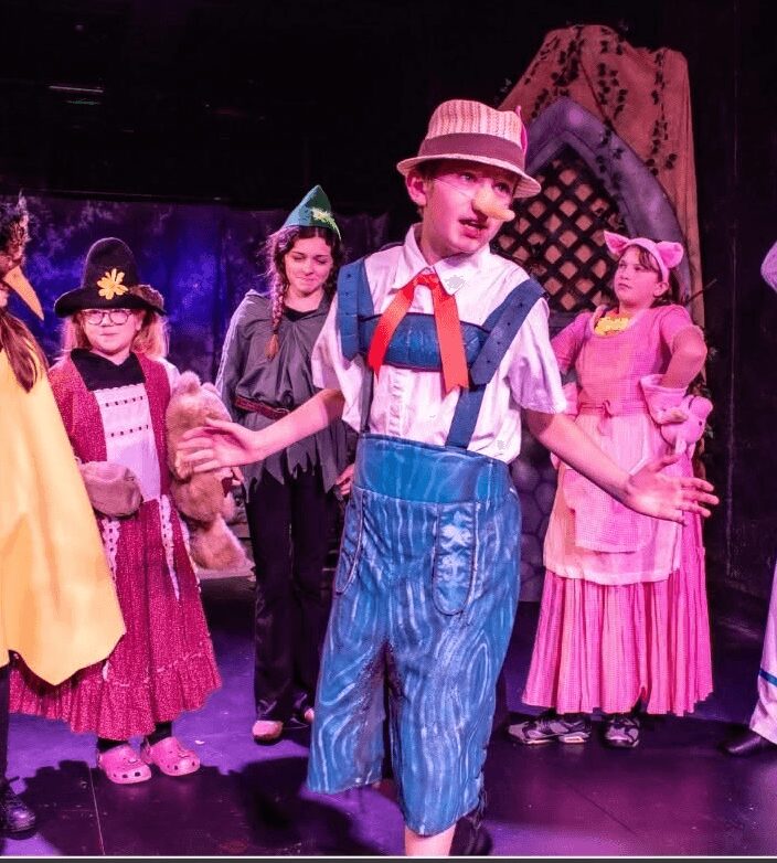 Children in colorful costumes perform on stage, with a boy in striped pants and a hat in the center.