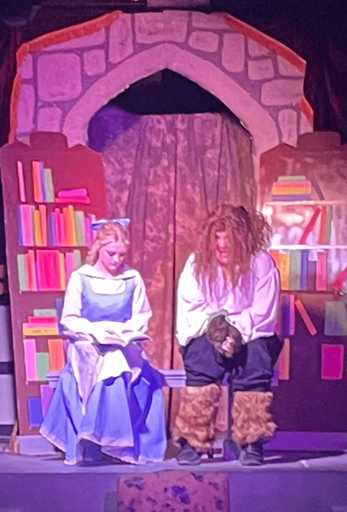 Two people on stage with a castle backdrop and bookshelves, one seated in a costume, the other standing with a dog.