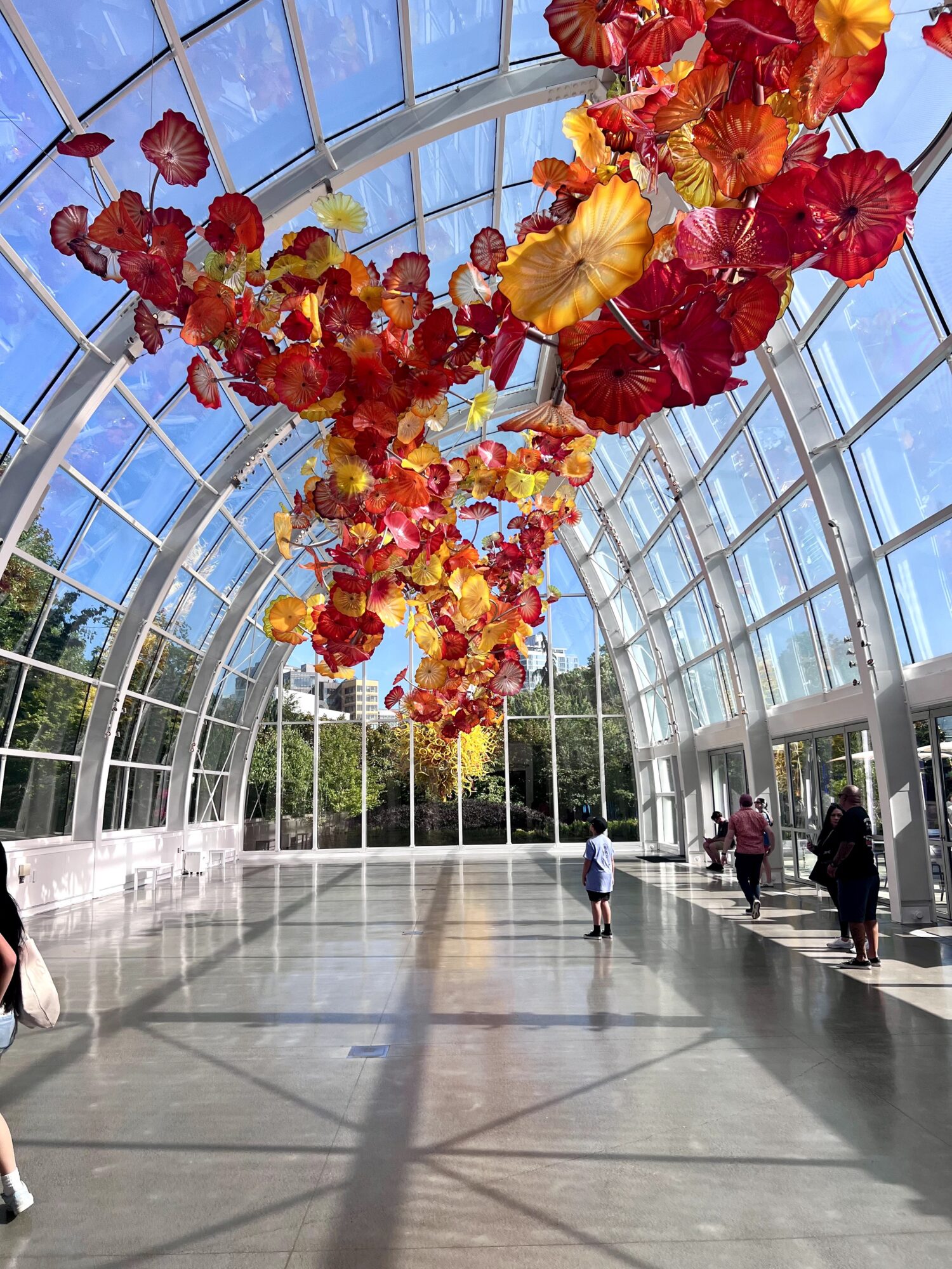 Colorful glass sculpture hanging inside a glass conservatory with people walking and sunlight streaming in.
