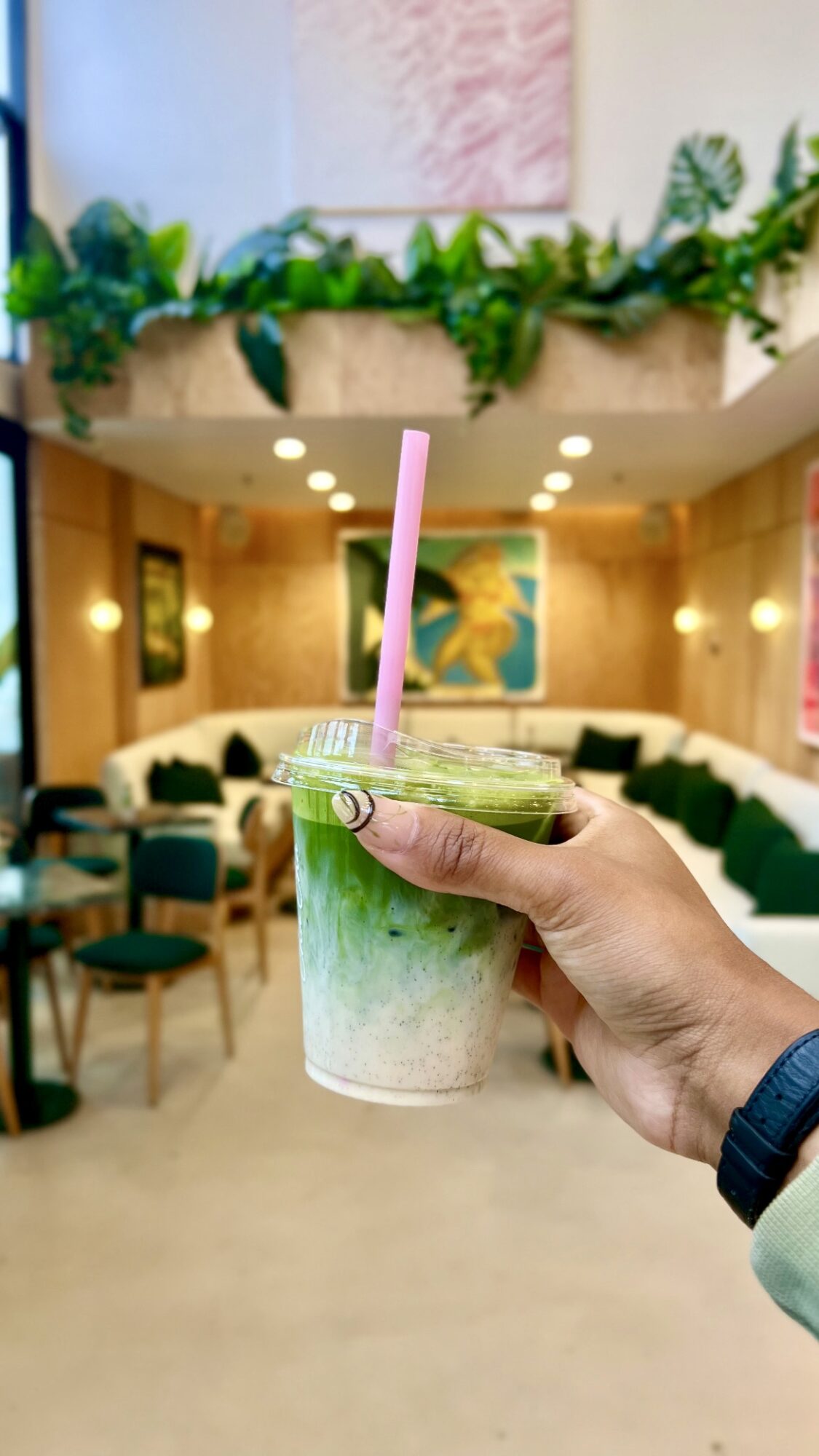 Hand holding a layered green and white drink with a pink straw in a cafe, with seating and artwork in the background.