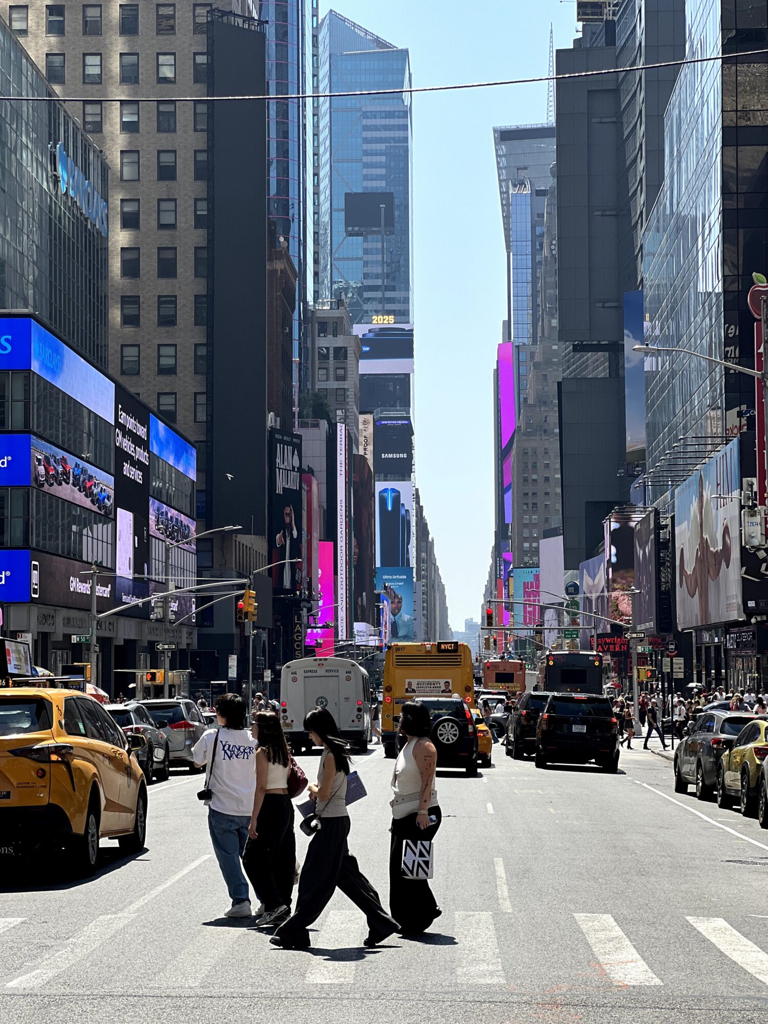 Street scene with pedestrians crossing, yellow taxis, and tall buildings with digital billboards in a city.