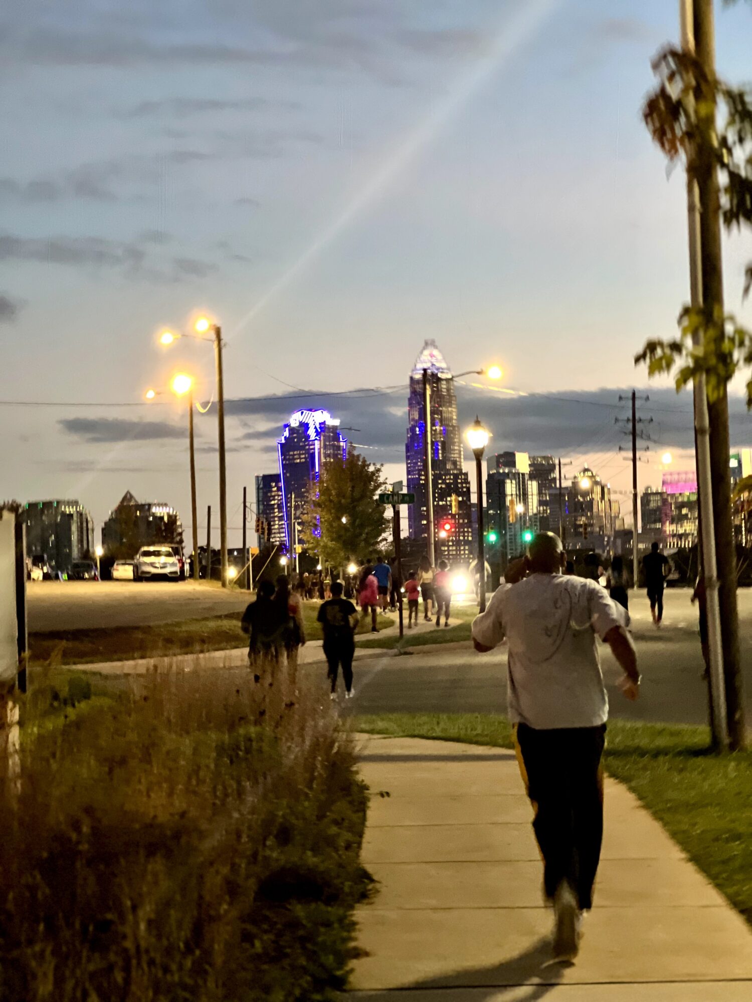 People walking on a city sidewalk with tall buildings and a rainbow in the sky, streetlights, and trees.