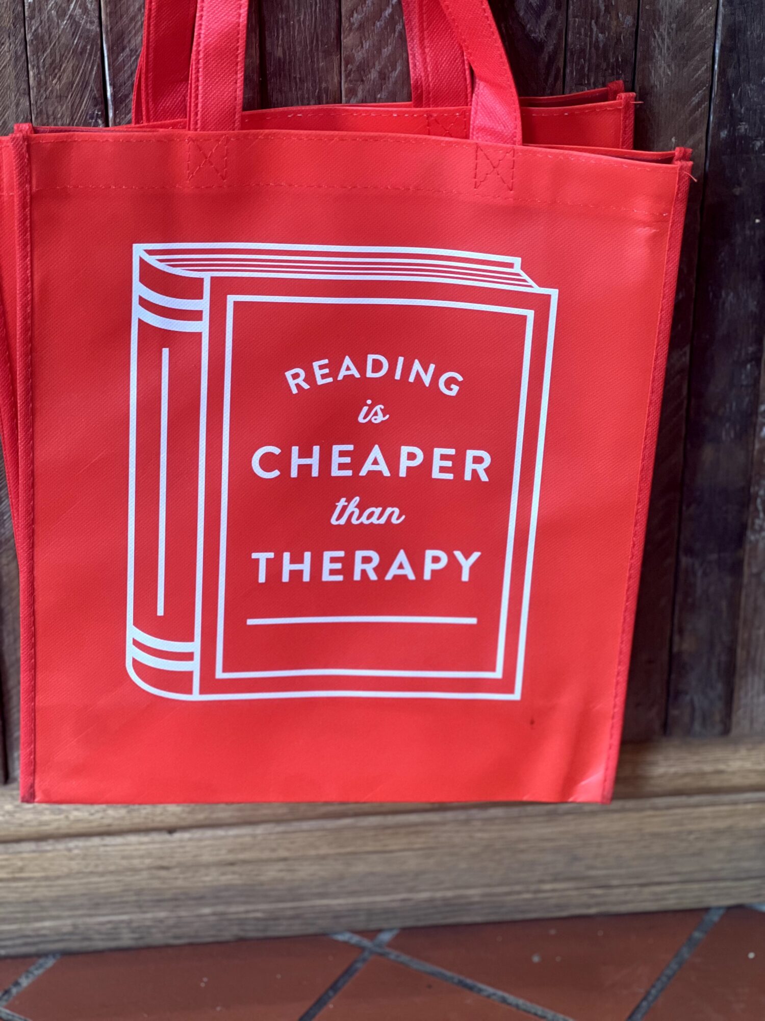 Red tote bag with white text reading 'Reading is Cheaper than Therapy' on a wooden surface.