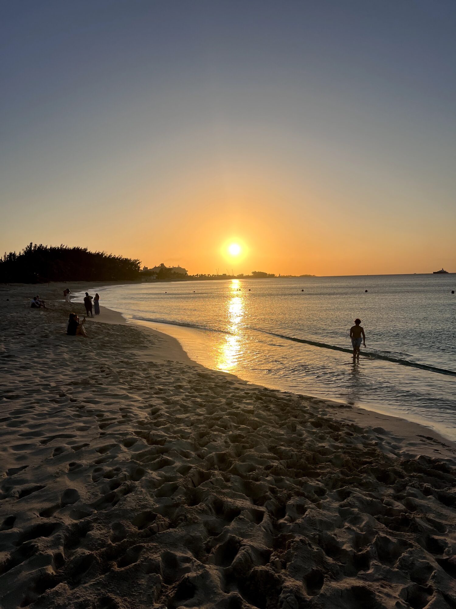 Sunset over a beach with people walking along the shoreline, calm water, and a distant tree line.