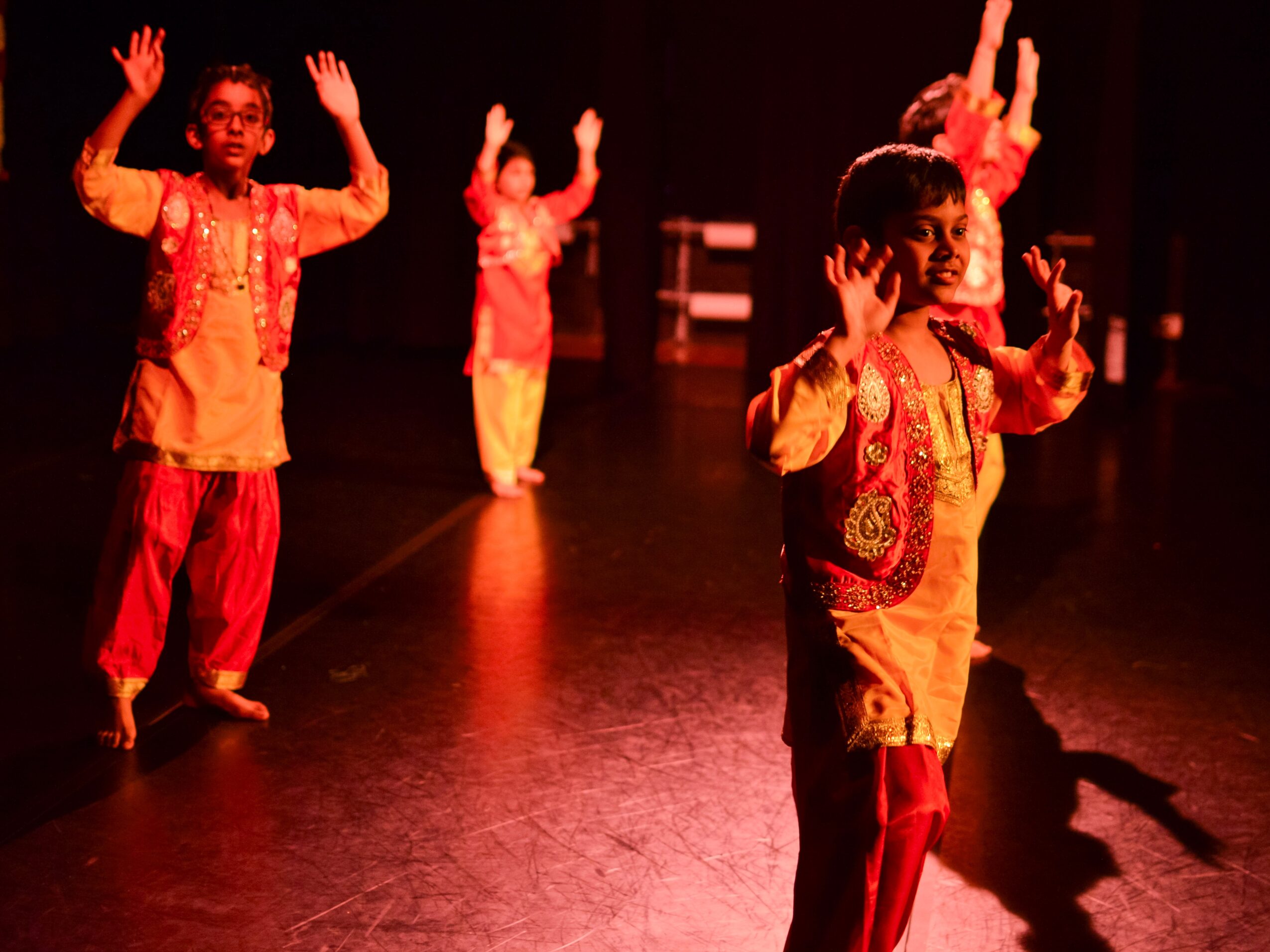 Children performing a dance on stage, wearing colorful traditional costumes, with arms raised and expressive poses.