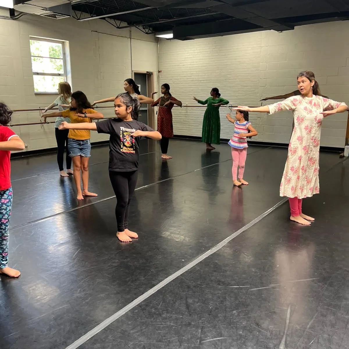 Group of children and an instructor practicing dance in a studio with black floor and white walls, arms extended outward.