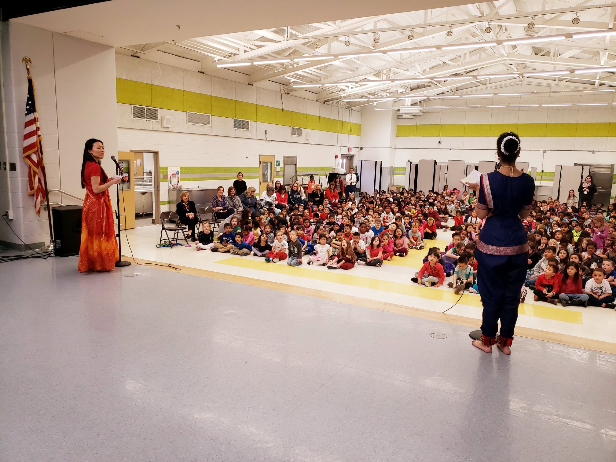 A woman in a red dress speaking on stage to a large group of children seated on the floor in a gymnasium, with another woman standing nearby.