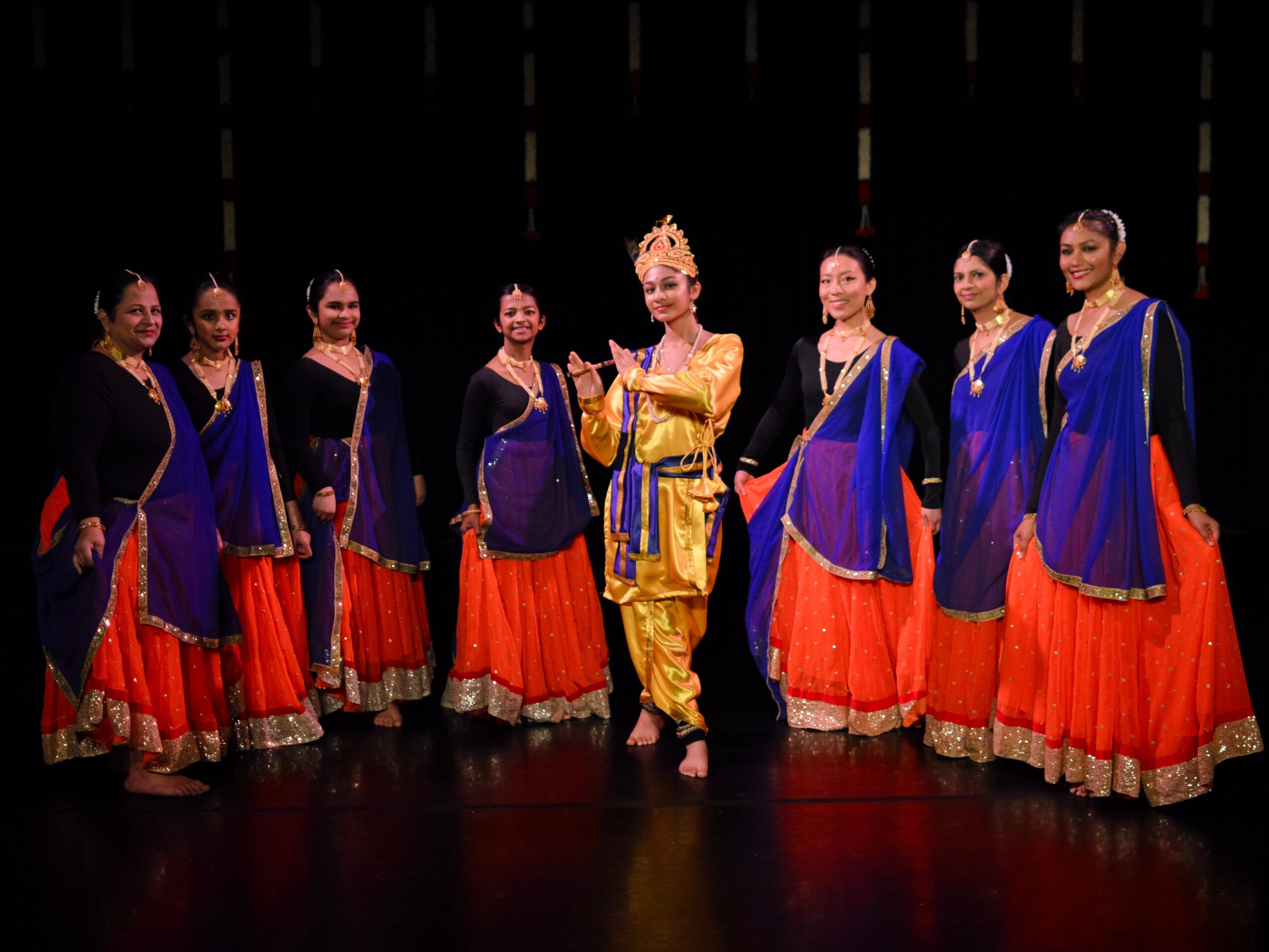 Group of seven women in colorful traditional costumes standing on stage, one in gold attire, others in blue and orange outfits.