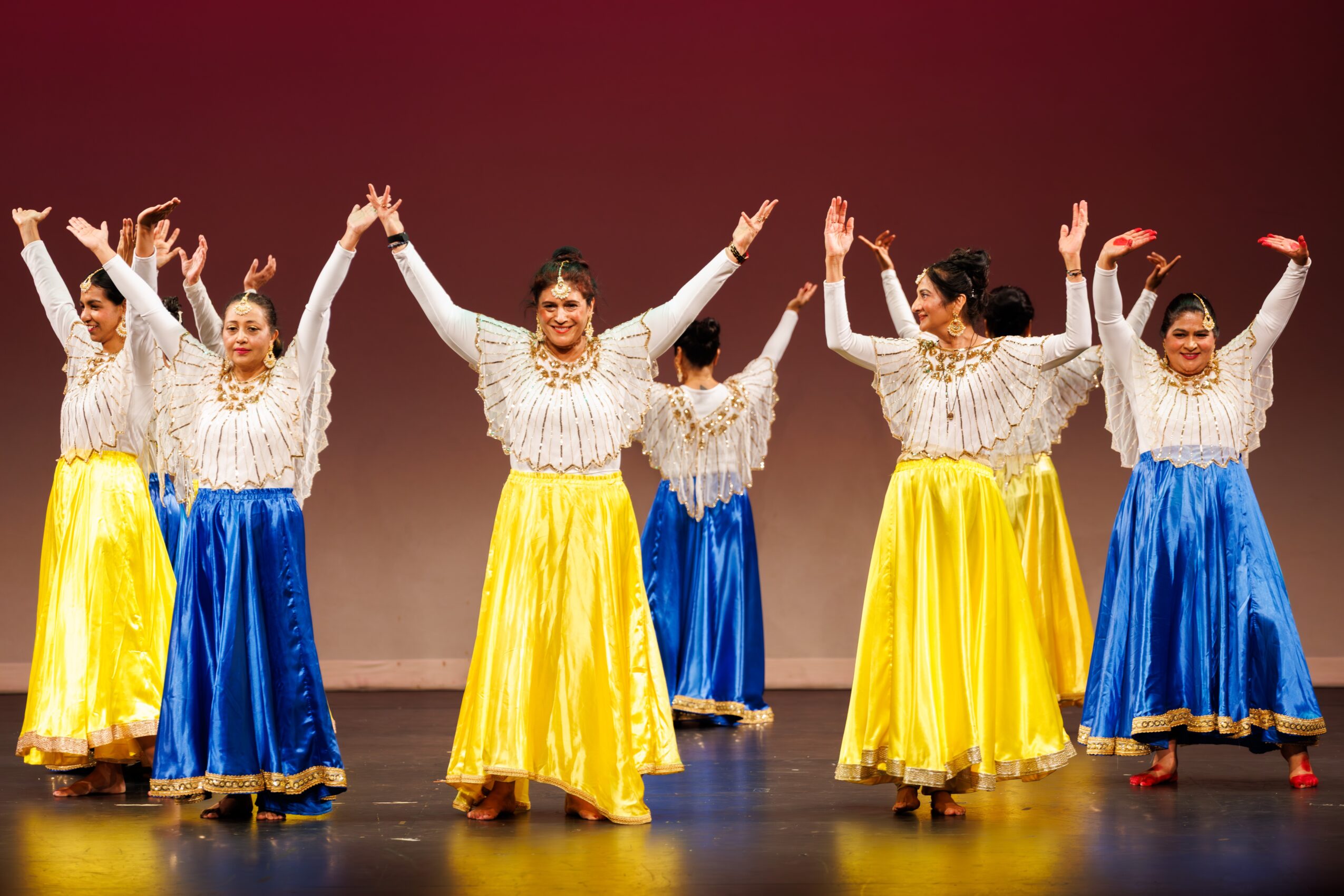 Group of six women dancing on stage, wearing white tops and yellow and blue skirts, with arms raised.