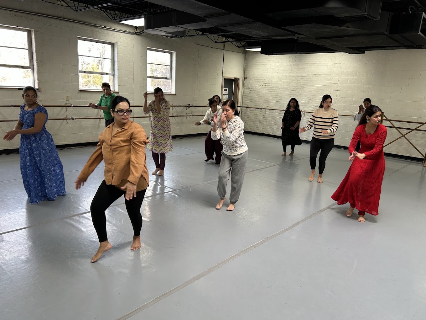 Group of women practicing dance in a studio with large windows and ballet barres, some standing and others moving.