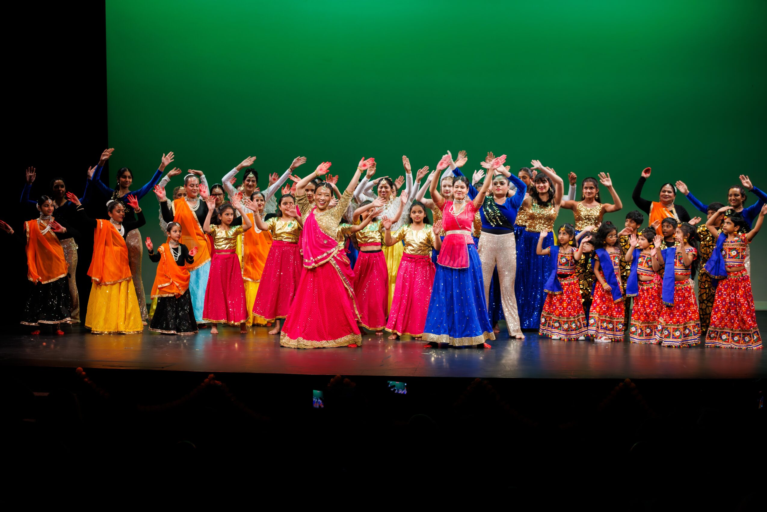 Group of children and adults in colorful costumes performing on stage with a green background.