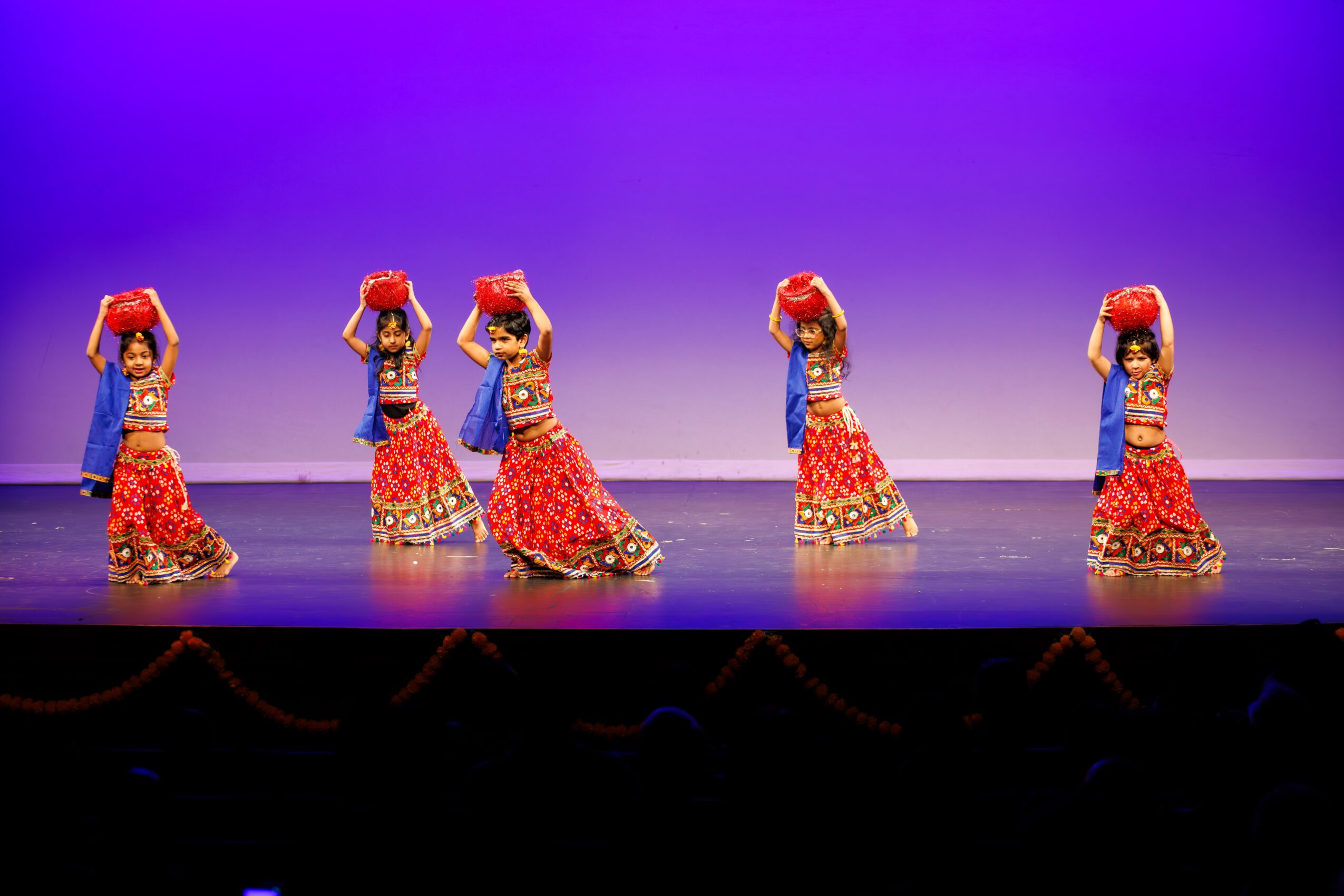 Five dancers in traditional costumes perform on stage with purple background, holding red objects above their heads.