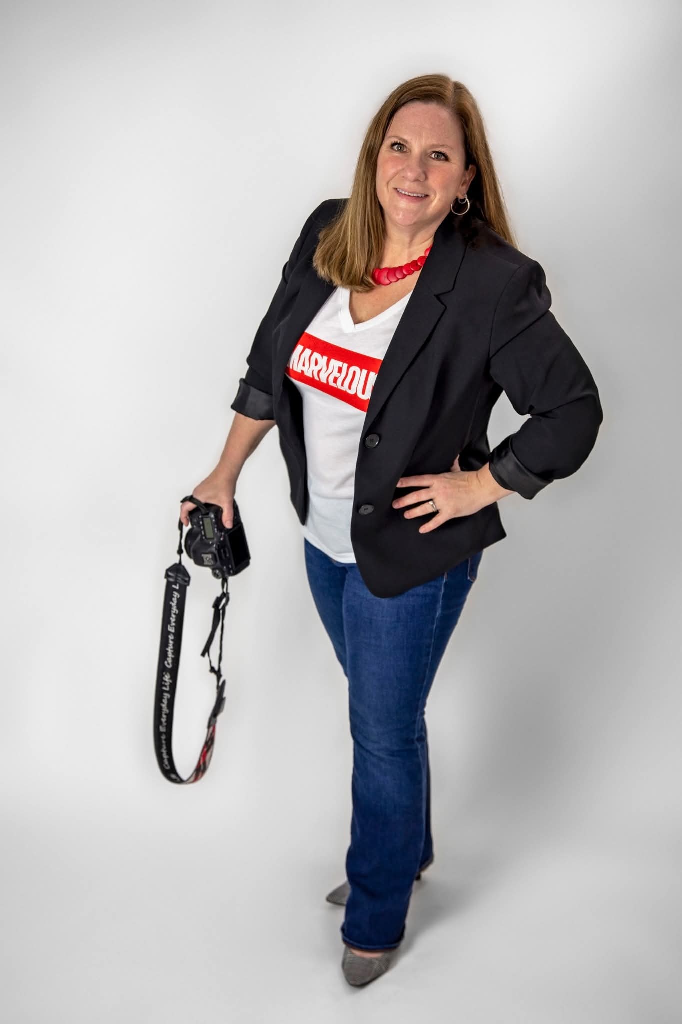 Woman standing with a camera in her right hand, smiling, wearing a black blazer, white t-shirt, and blue jeans, against a white background.