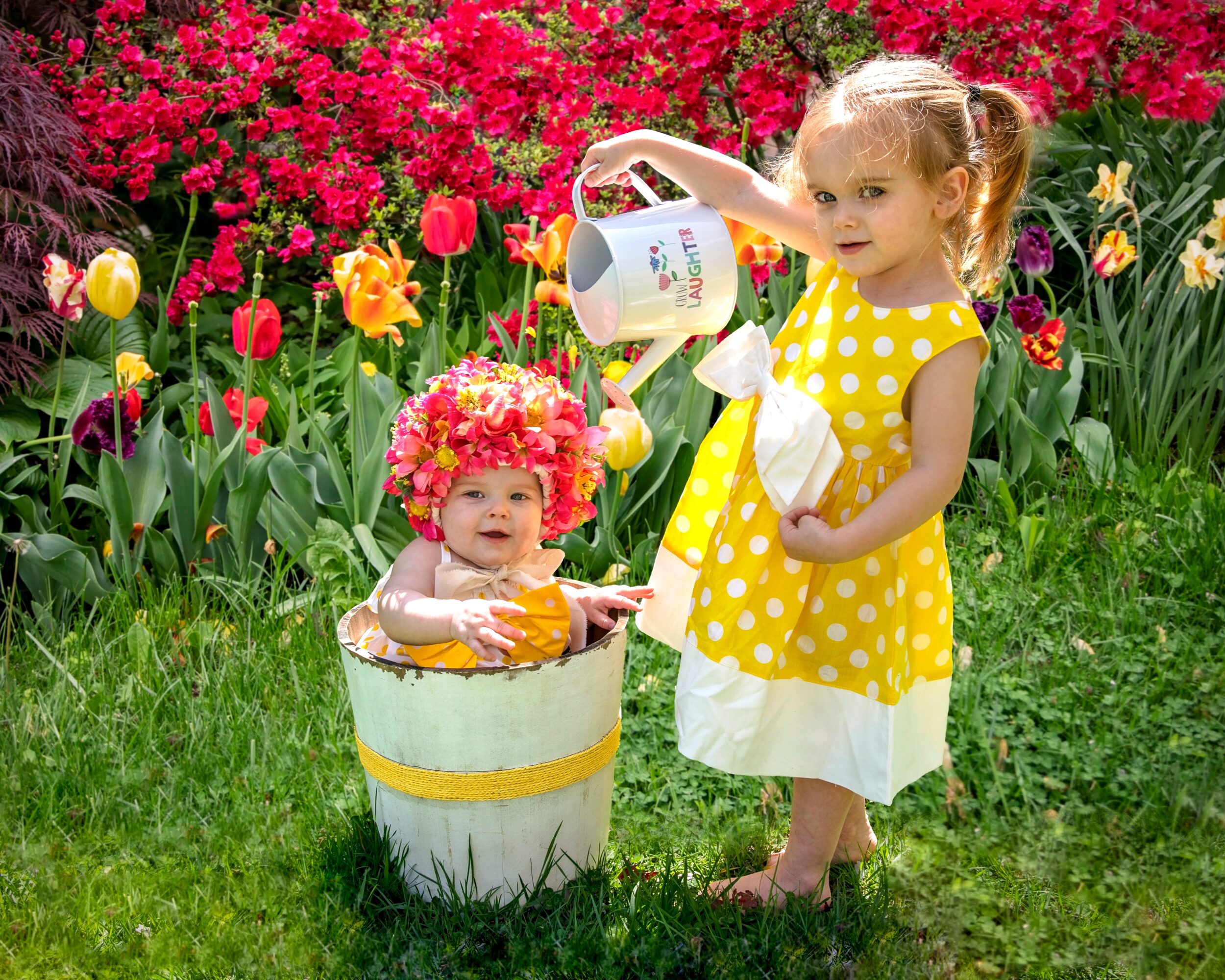Two young girls in a garden with colorful flowers, one watering the other in a bucket, wearing bright dresses.