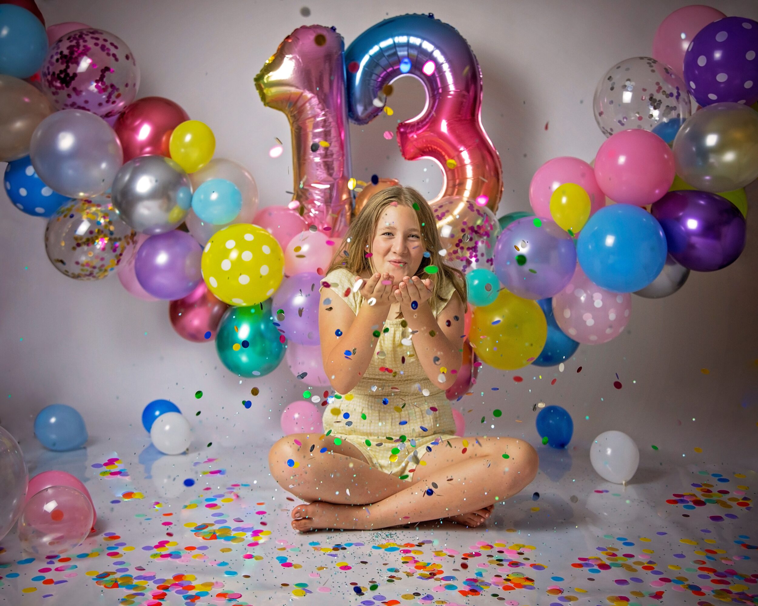 Girl sitting on floor with balloons and confetti, celebrating her 13th birthday, smiling with hands near face.
