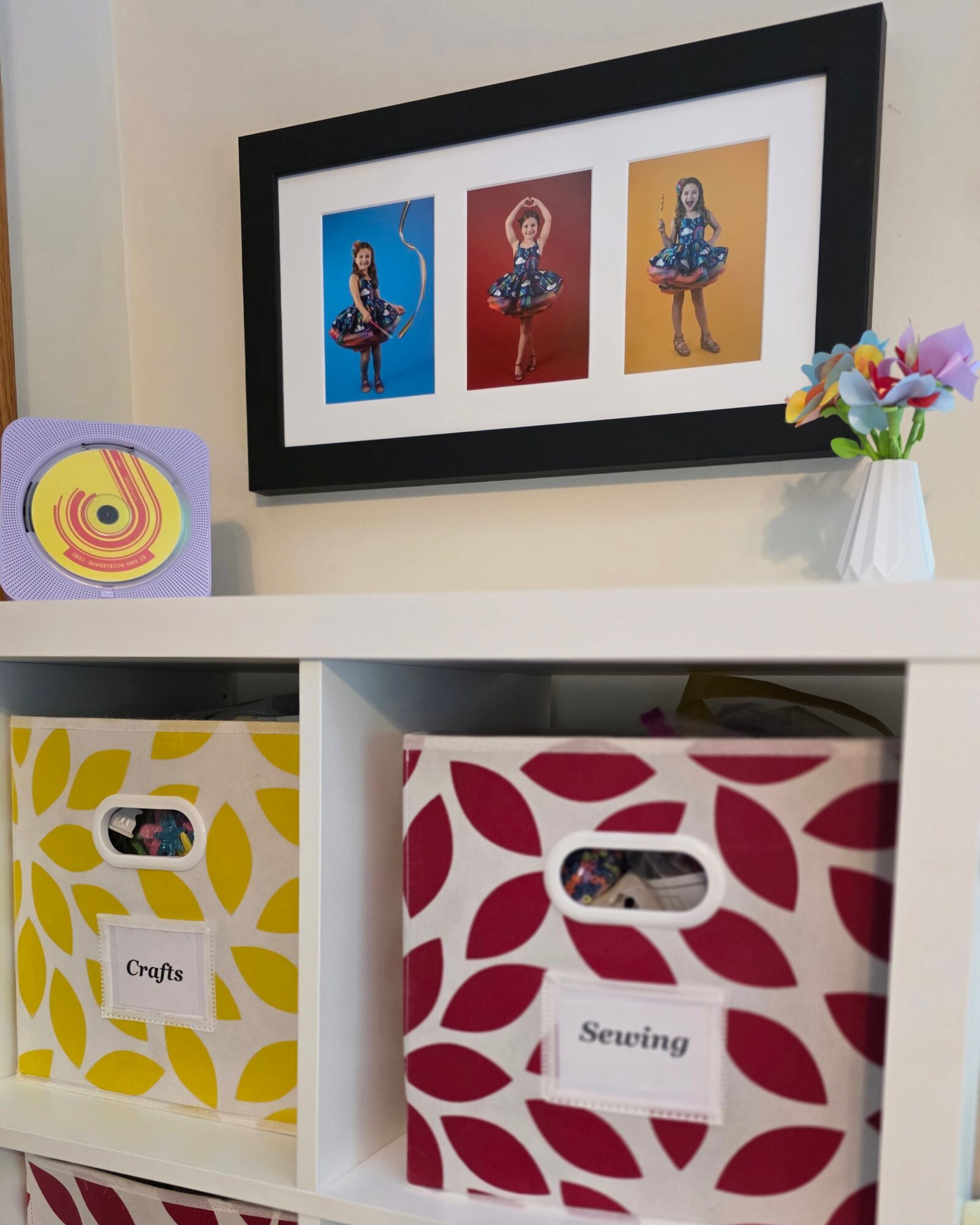 Framed artwork of three colorful figures hangs above a white shelf with decorative boxes and a flower vase.