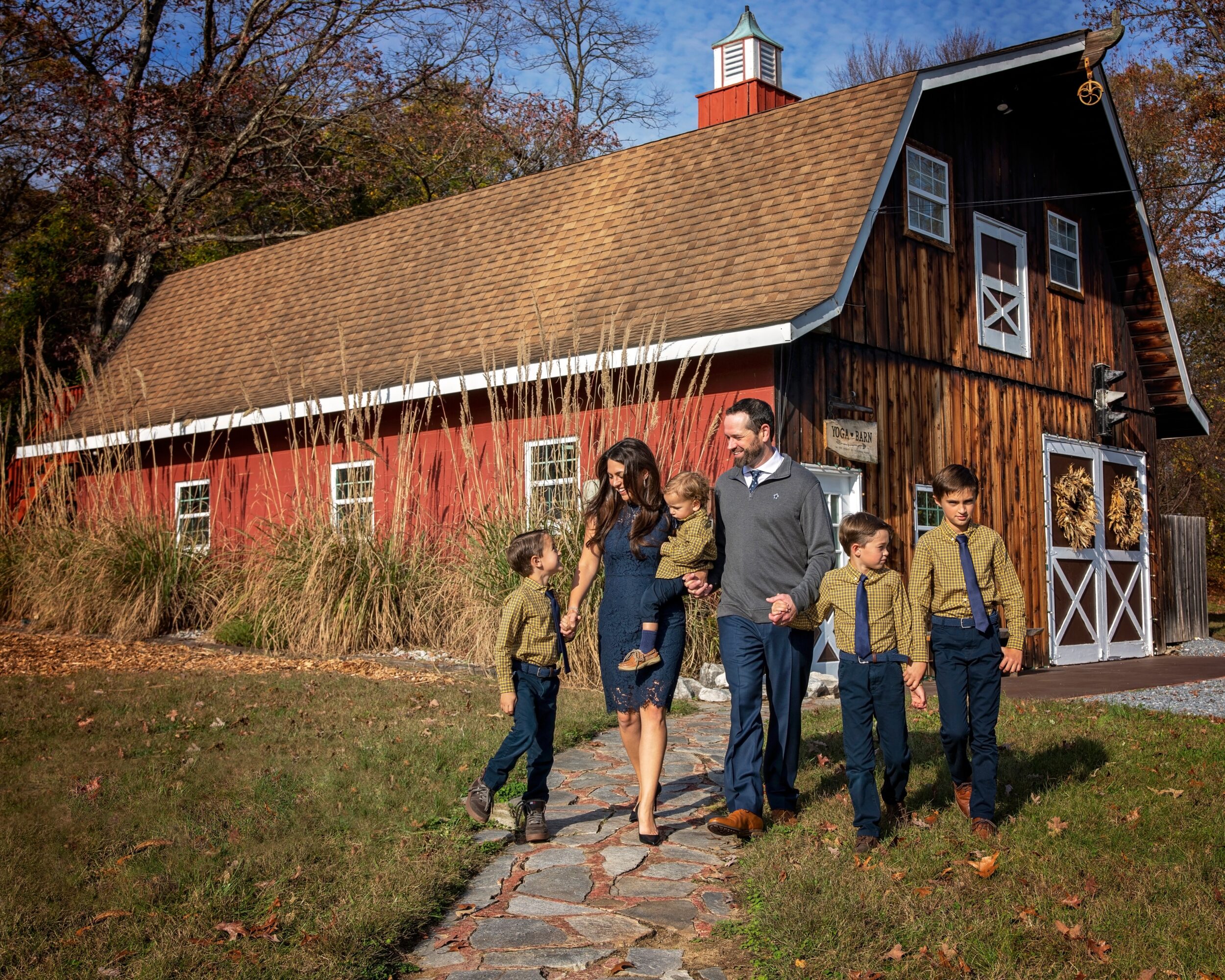 A family of six walking on a stone path in front of a barn with a brown roof and red and dark wood walls, trees in background.