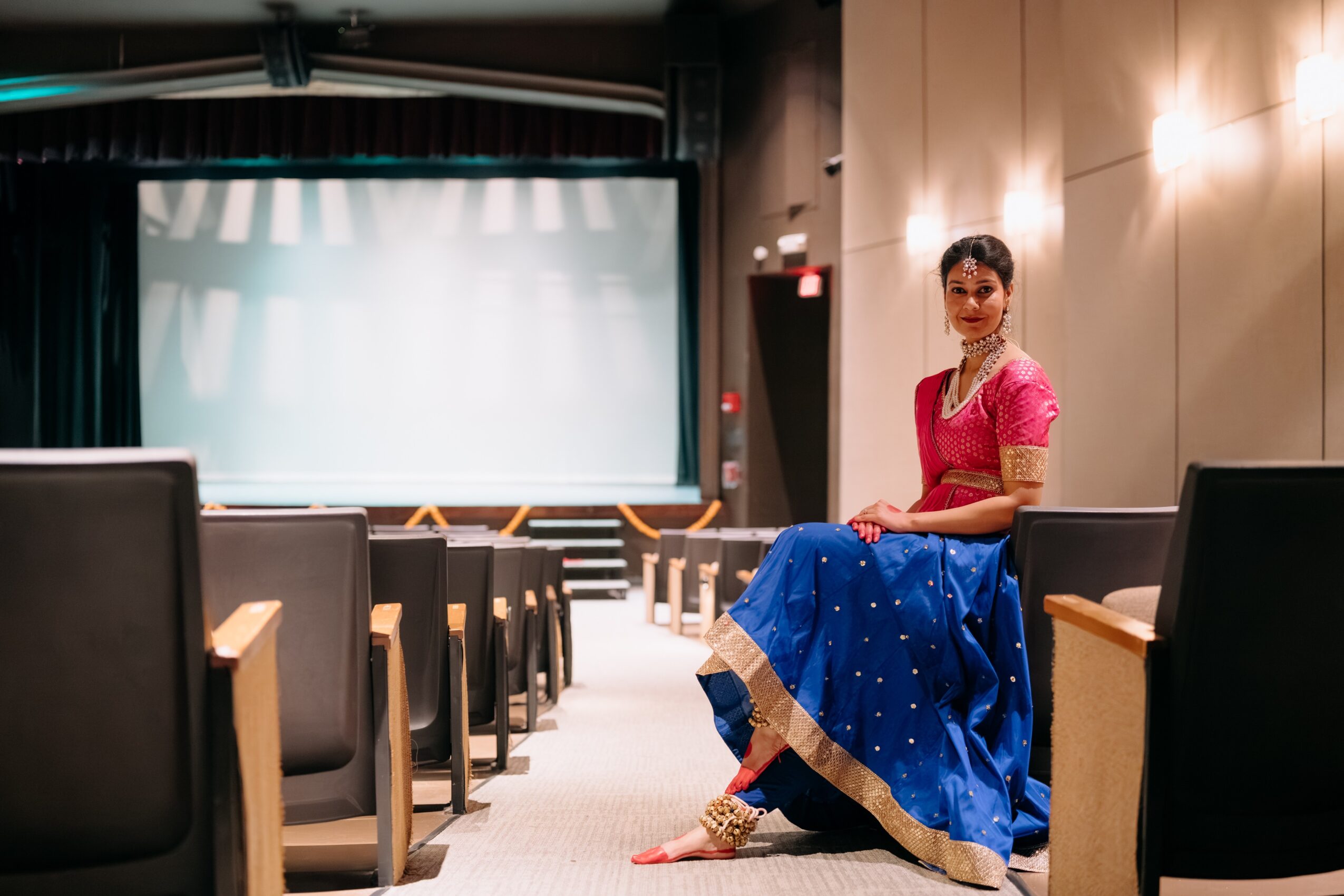 Woman in pink top and blue skirt sitting in an empty auditorium with chairs and a large screen behind her.