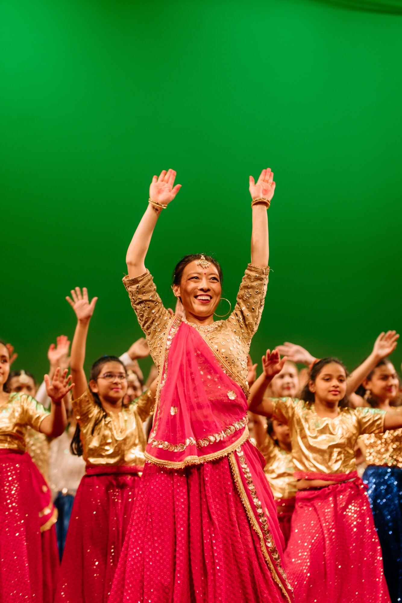 Group of children and a woman in colorful traditional clothing, smiling and raising hands, against a green background.