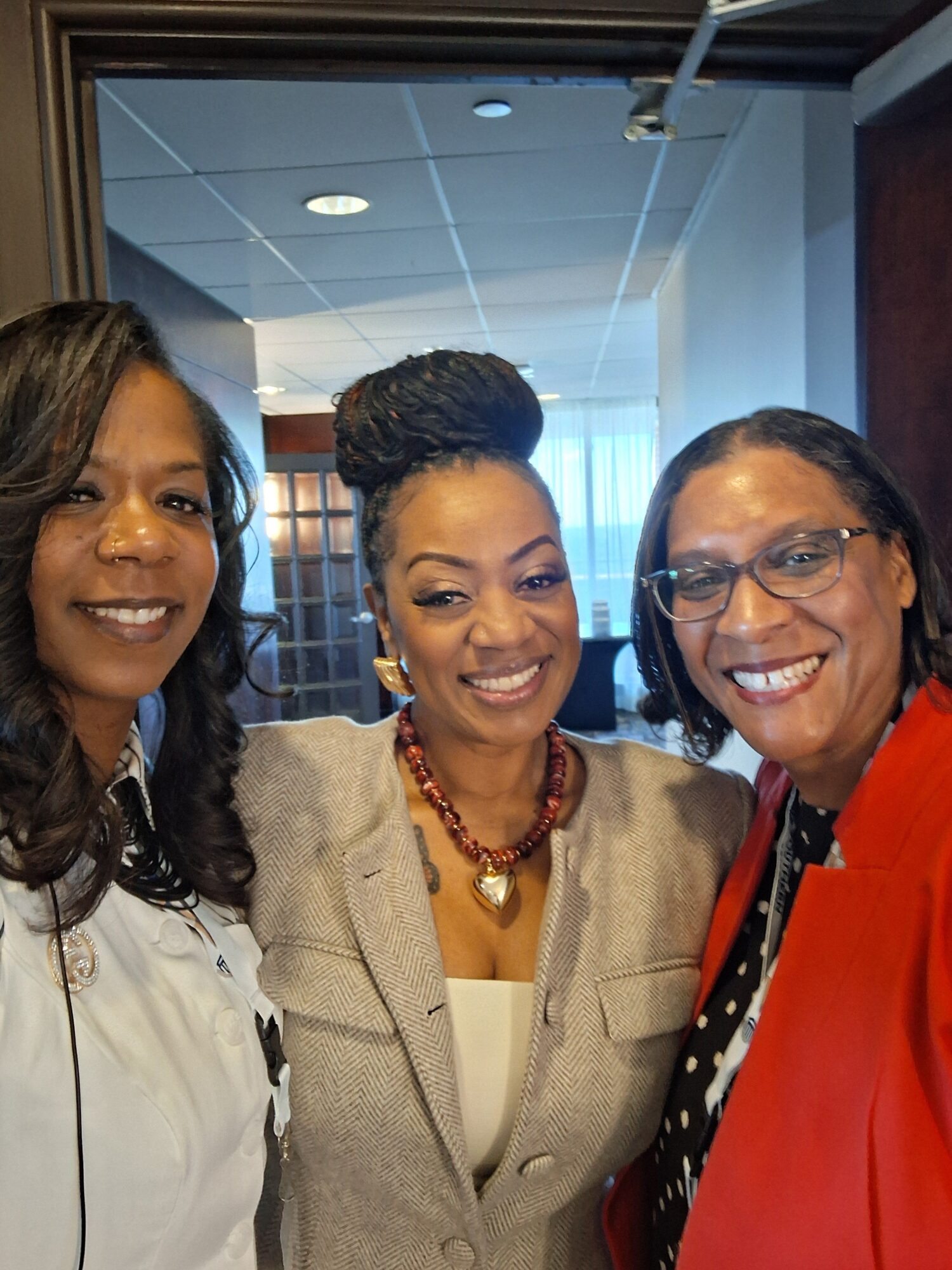 Three women smiling indoors, two wearing glasses, one with braided hair, all dressed professionally.