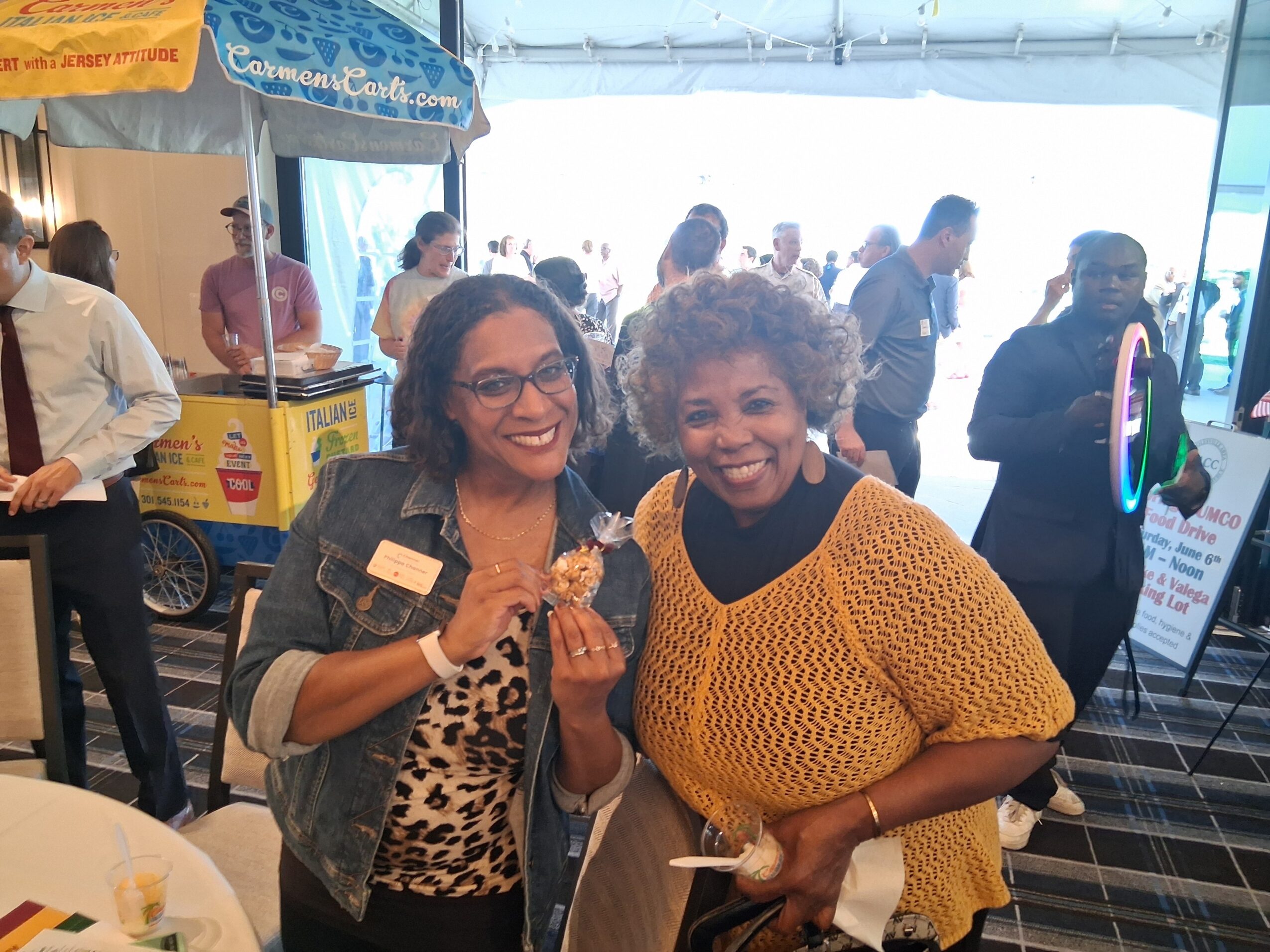 Two women smiling and holding food at an indoor event with people and booths in the background.