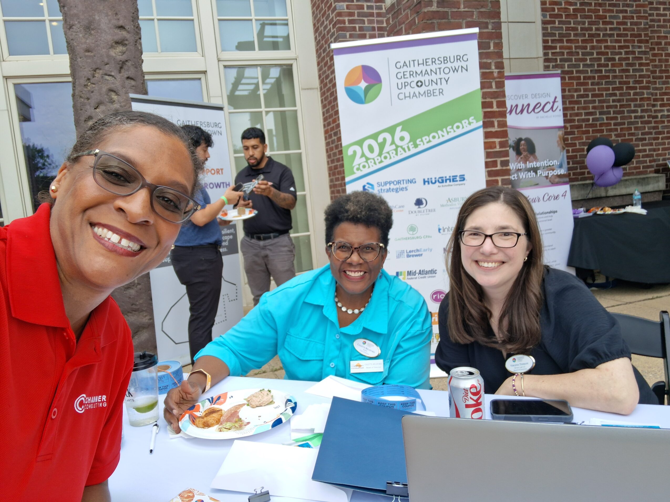 Three women smiling at an outdoor event, with a banner and tables in the background.