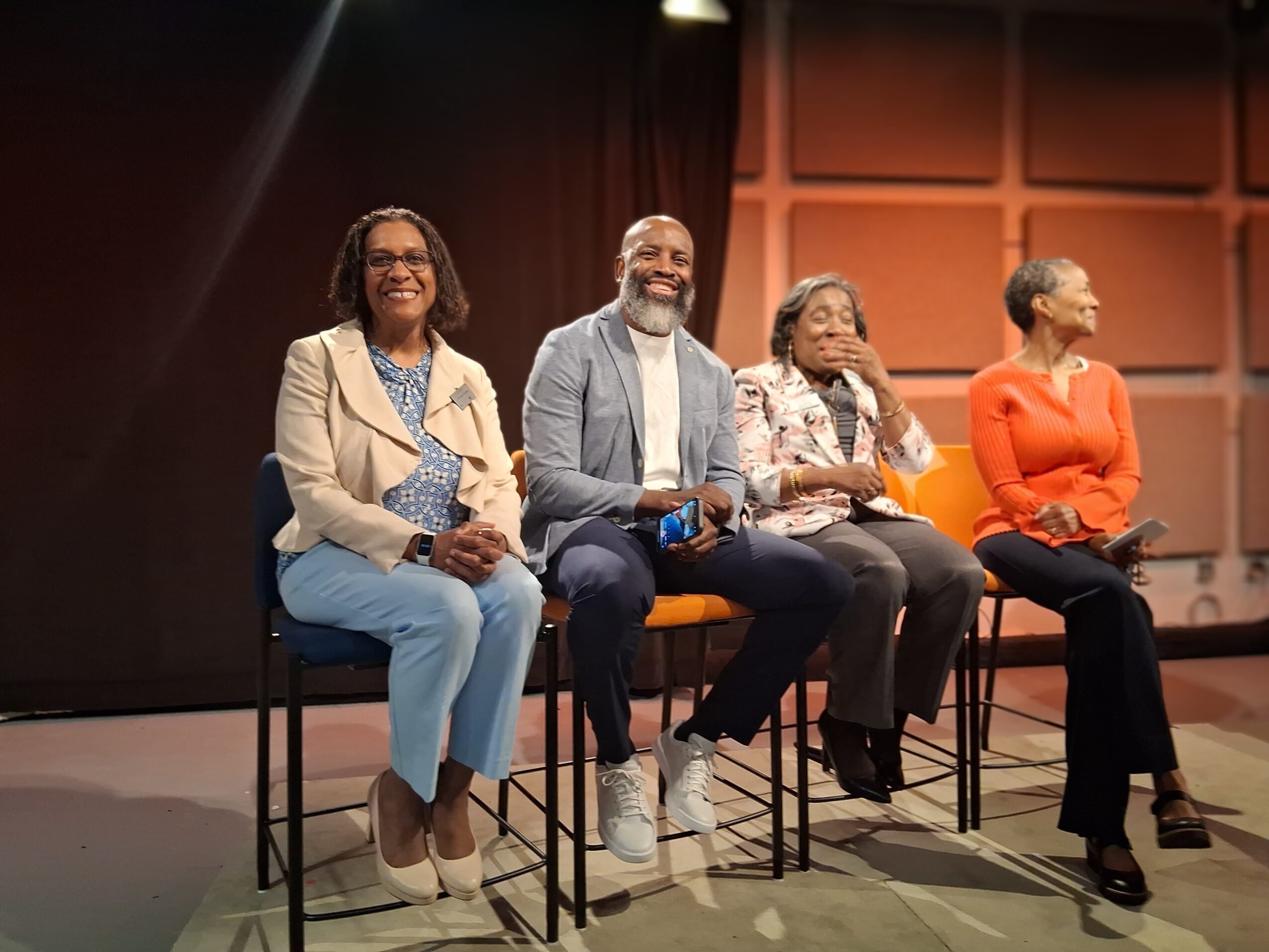 Four people sitting on chairs on stage, smiling and engaging with an audience, with a dark background and warm lighting.