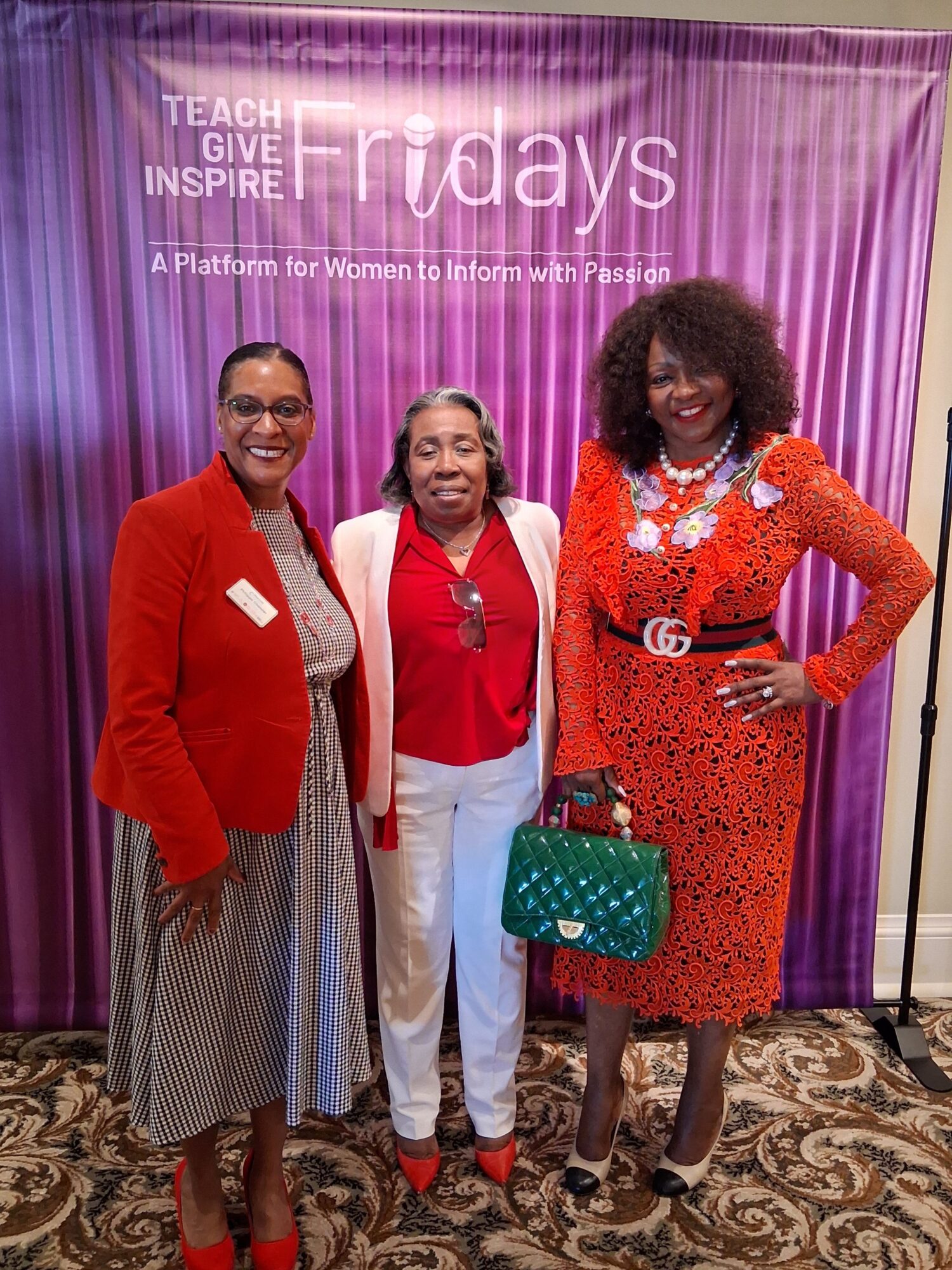 Three women standing in front of a purple curtain, smiling, with one holding a green purse, all dressed in colorful outfits.