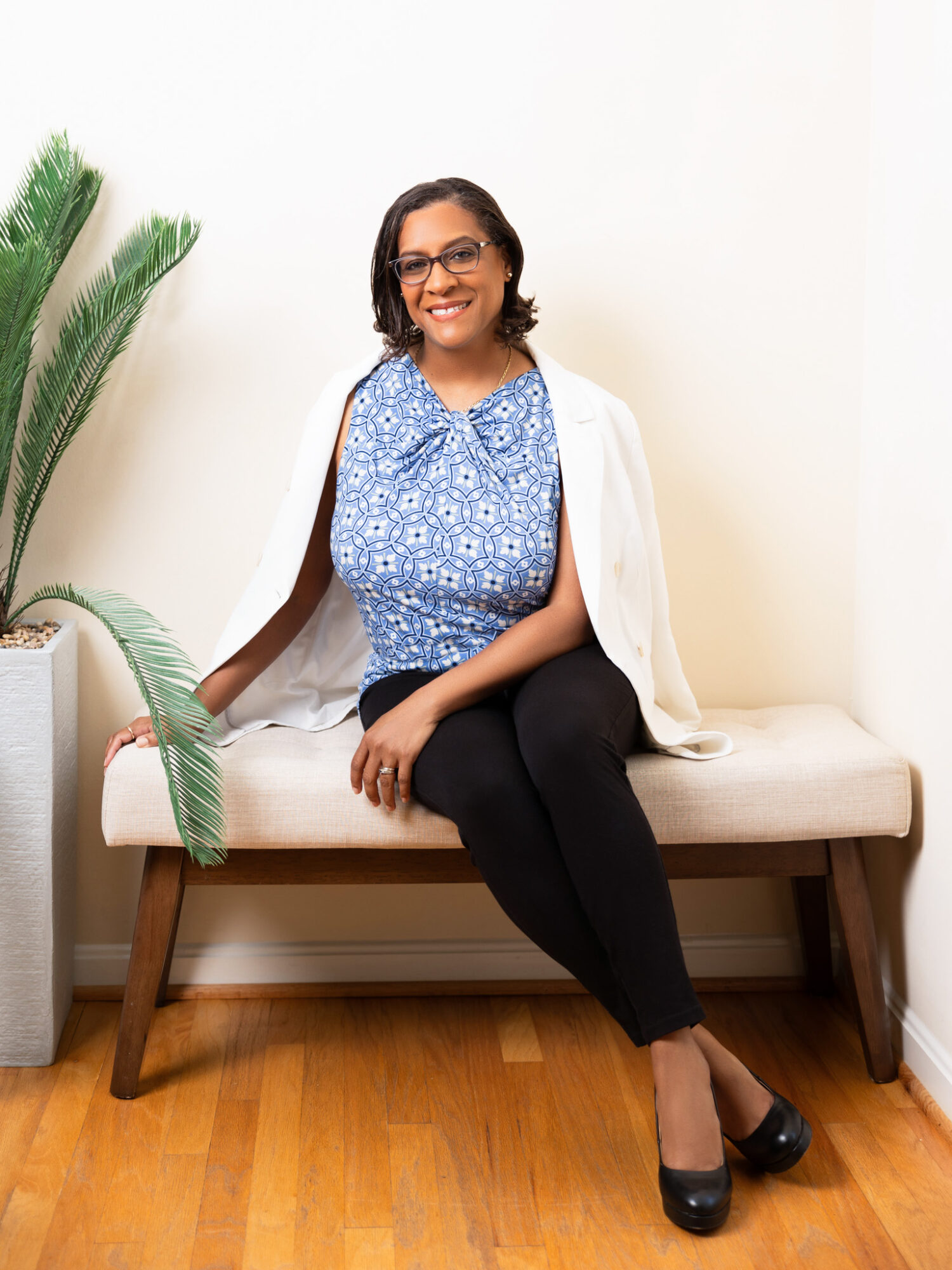 Woman sitting on a bench, smiling, wearing glasses, a blue patterned blouse, black pants, and black shoes, with a plant nearby.