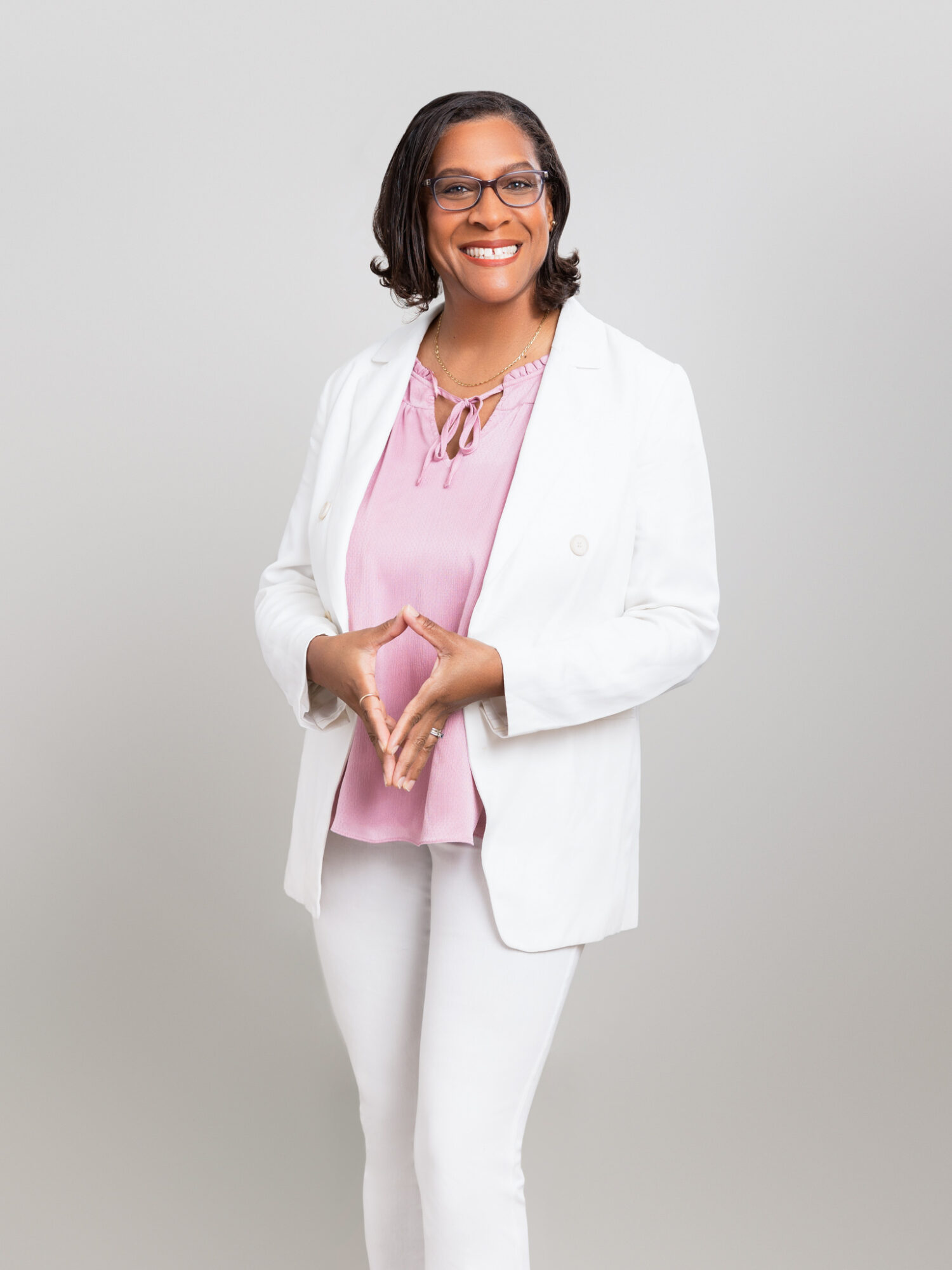 Smiling woman with glasses in a white blazer and pink blouse, standing against a plain light background.