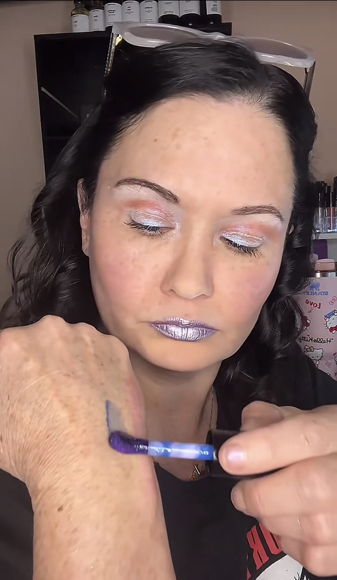 Person with makeup applying eyeshadow and lipstick, holding a makeup brush, in a room with shelves in the background.