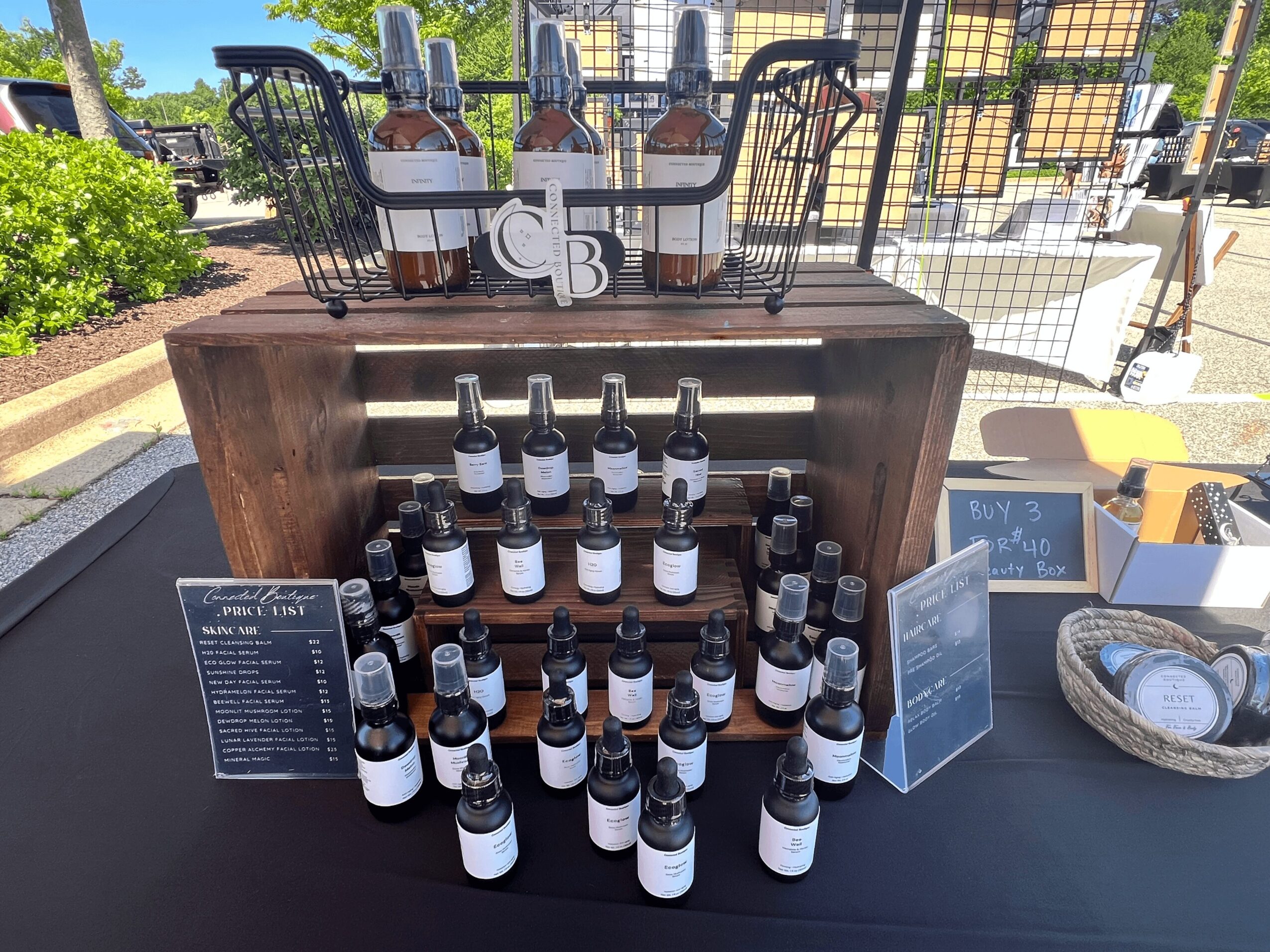 Display of multiple bottles on a table with a wooden box, outdoor setting, and signs, possibly at a market or event.