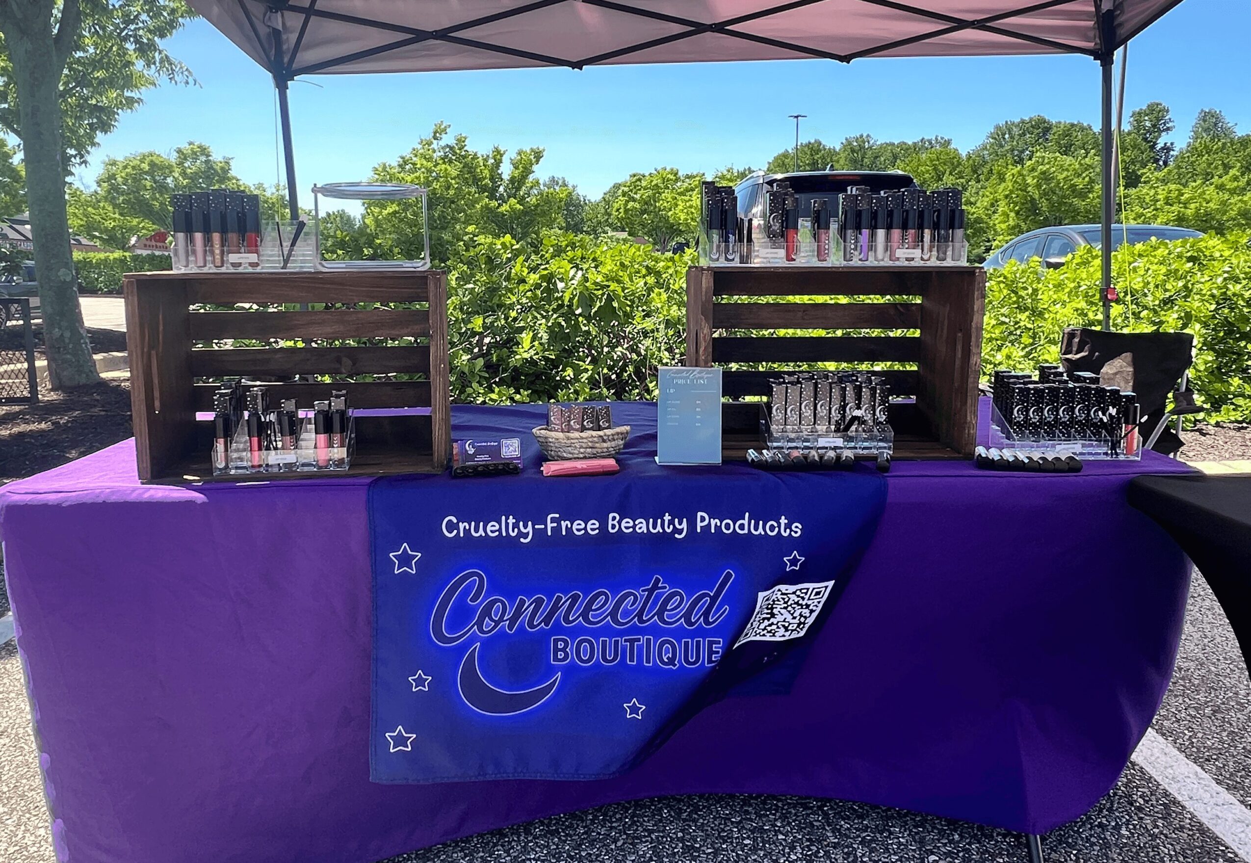 Outdoor booth with purple tablecloth displaying cruelty-free products under a canopy, with trees and parking lot in background.
