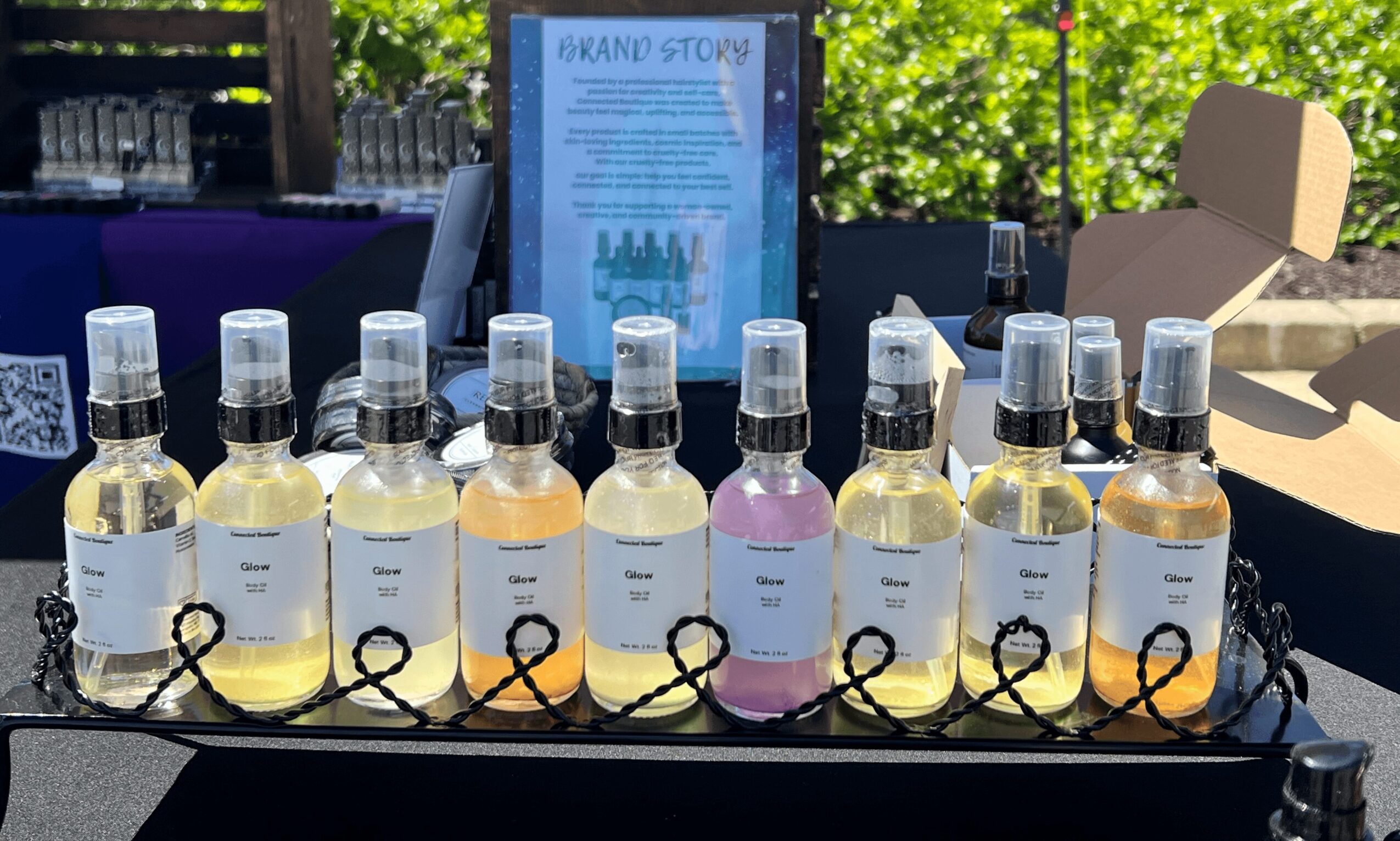 Display of ten bottles with clear liquid, labels, and spray caps, arranged on a table outdoors with trees and a sign in the background.