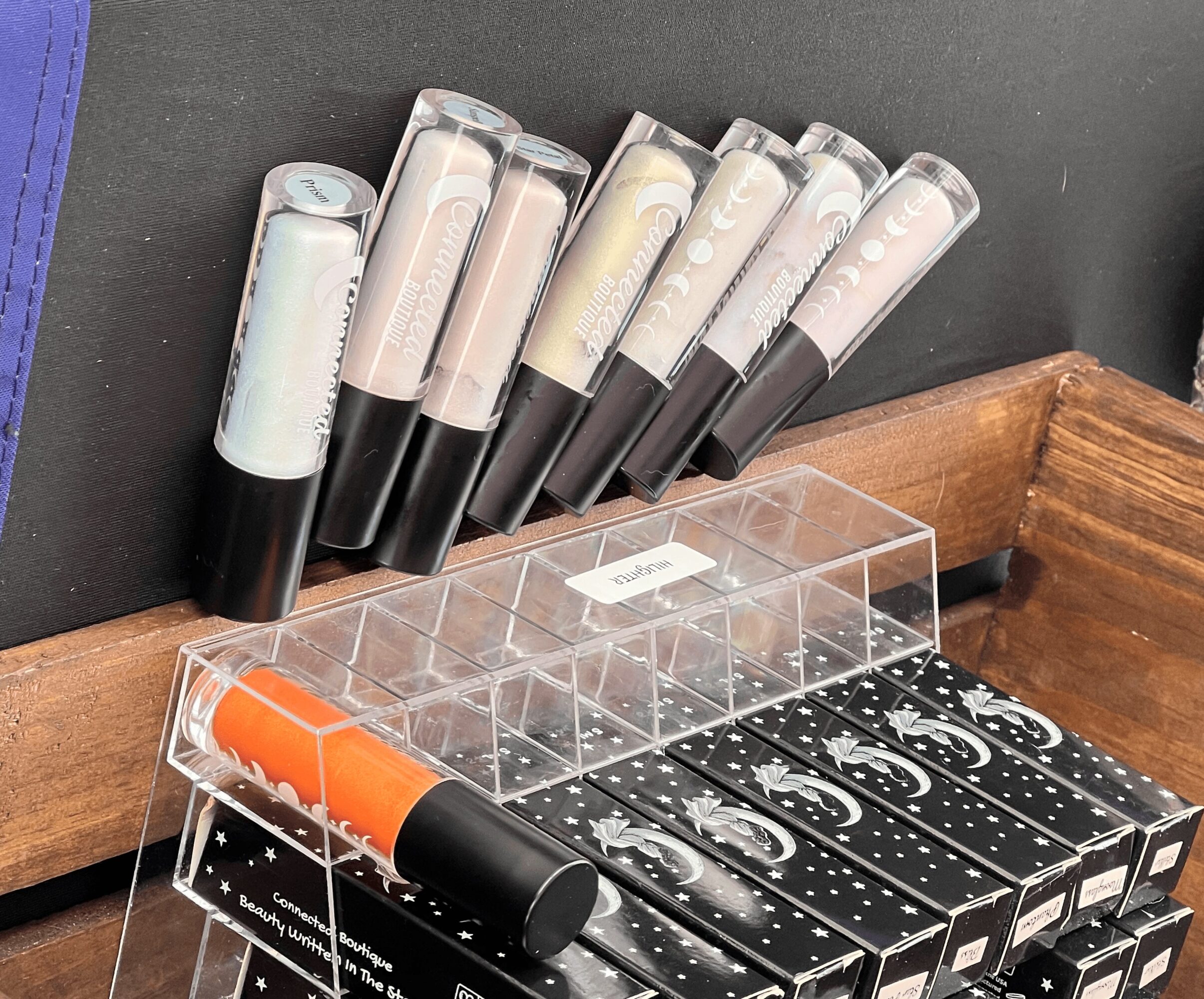 Six lip gloss tubes with black caps arranged on a black surface near a wooden edge and a clear plastic container.