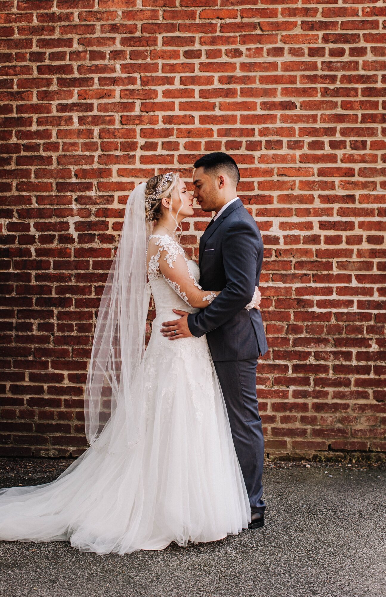Bride and groom face each other in front of a brick wall, holding hands, dressed in wedding attire.