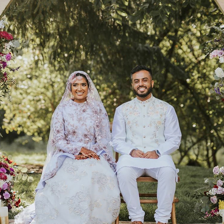 A bride and groom sit outdoors on a sunny day, surrounded by flowers and greenery, smiling at the camera.
