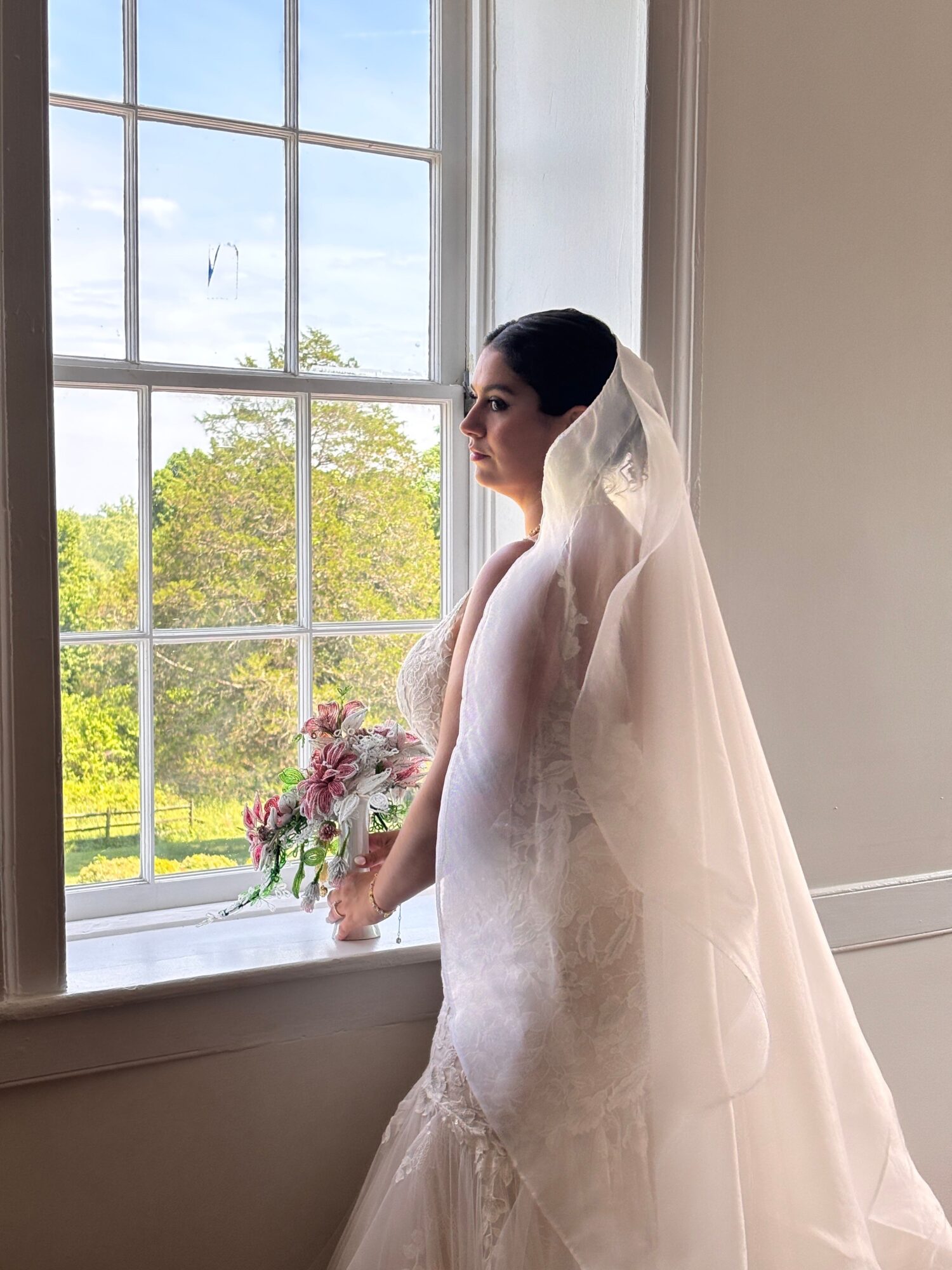 Woman in wedding dress holding bouquet, standing by window with outdoor view, sunlight streaming in, greenery outside.