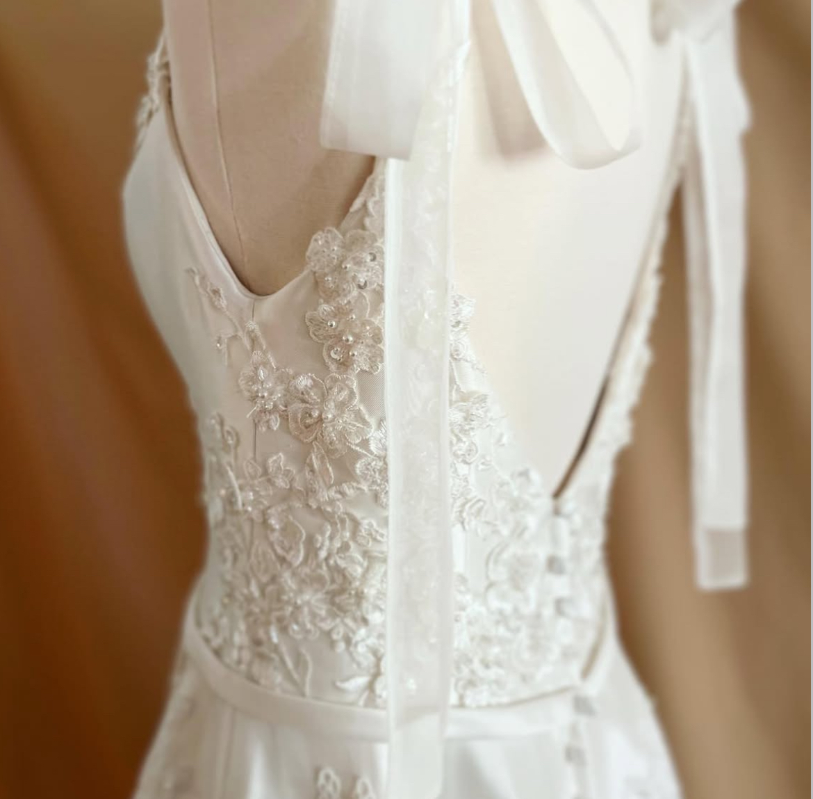 Close-up of a wedding dress with lace and embroidery, featuring a bow on the shoulder strap, against a wooden background.