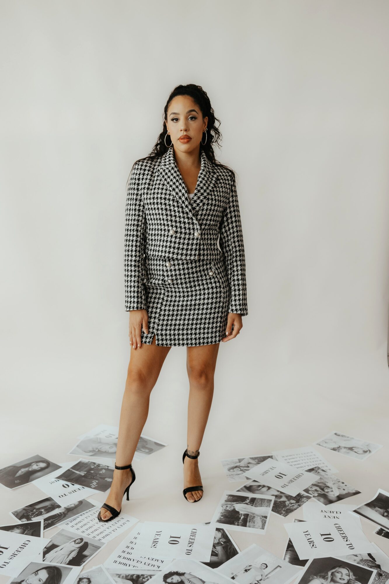 Woman in checkered blazer and skirt stands on scattered papers against plain background.