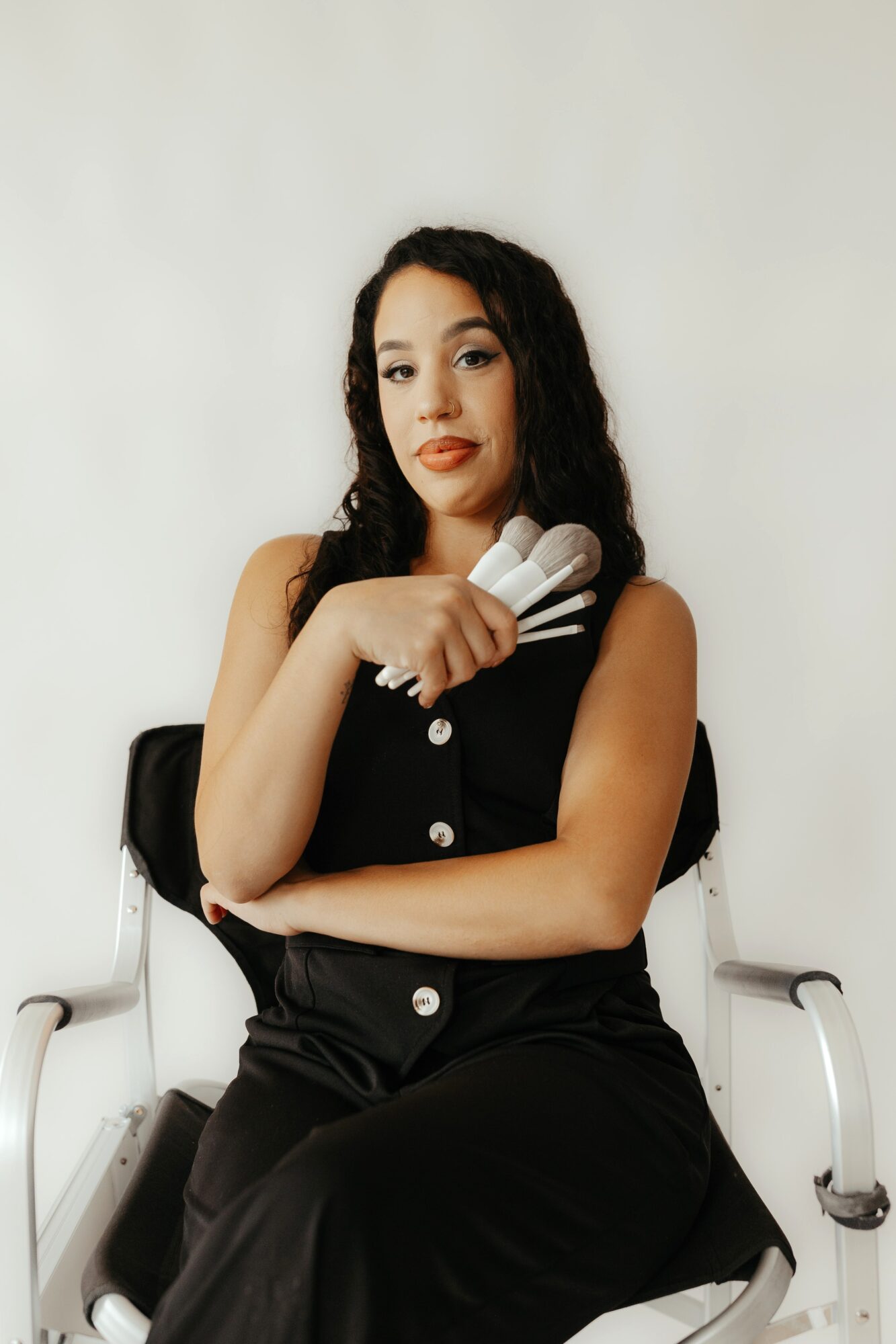 Young woman with curly hair sitting on a chair, holding a rolled-up towel, wearing a black sleeveless outfit, against a plain background.
