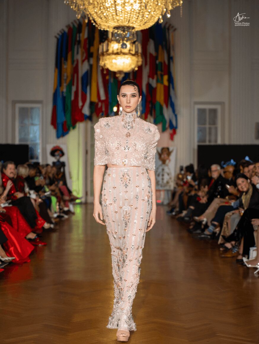 Model walking down runway in ornate dress with flags and chandelier in background, audience on both sides, hardwood floor.