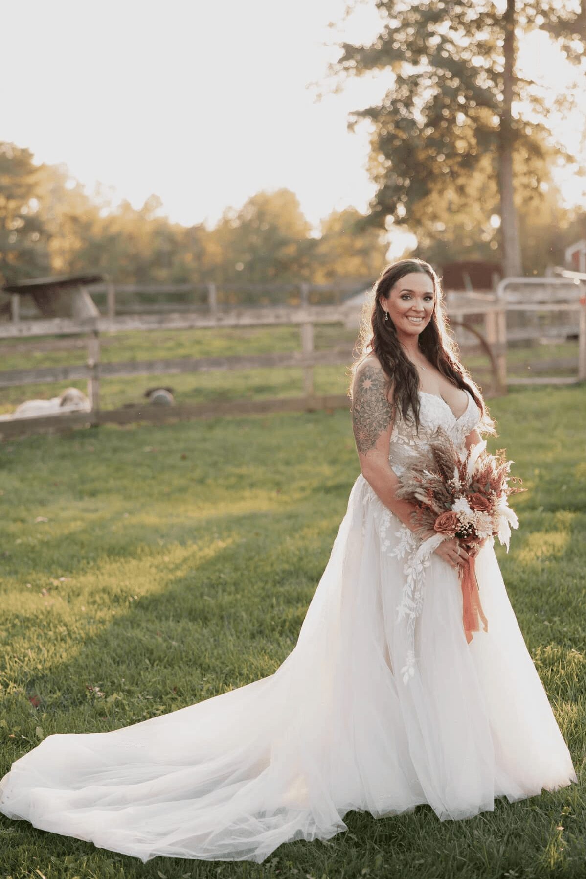 Woman in wedding dress holding a bouquet outdoors, smiling, with trees and a fence in the background.