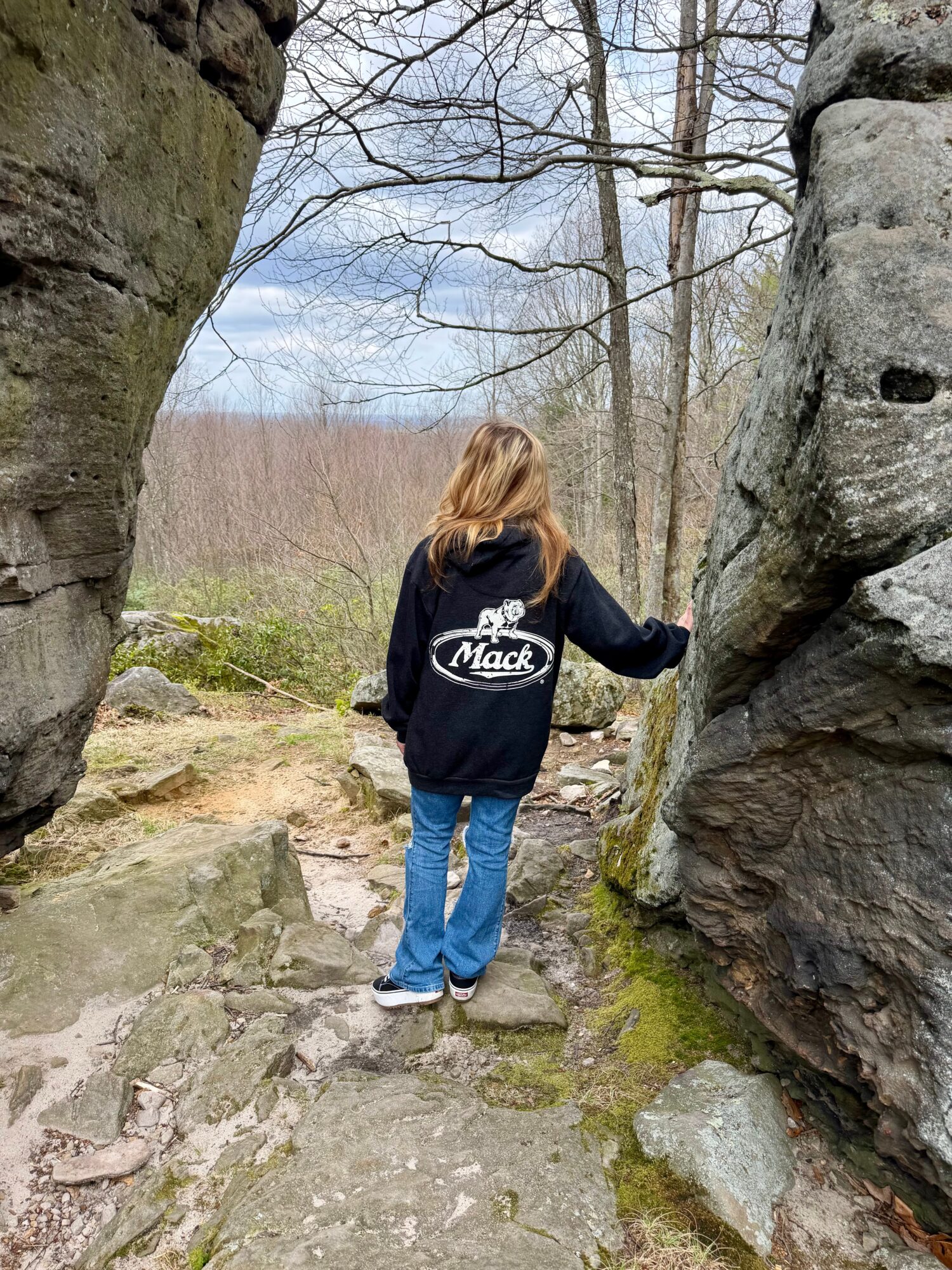 Person with long hair wearing a black jacket and blue jeans standing between large rocks outdoors, facing away from the camera.