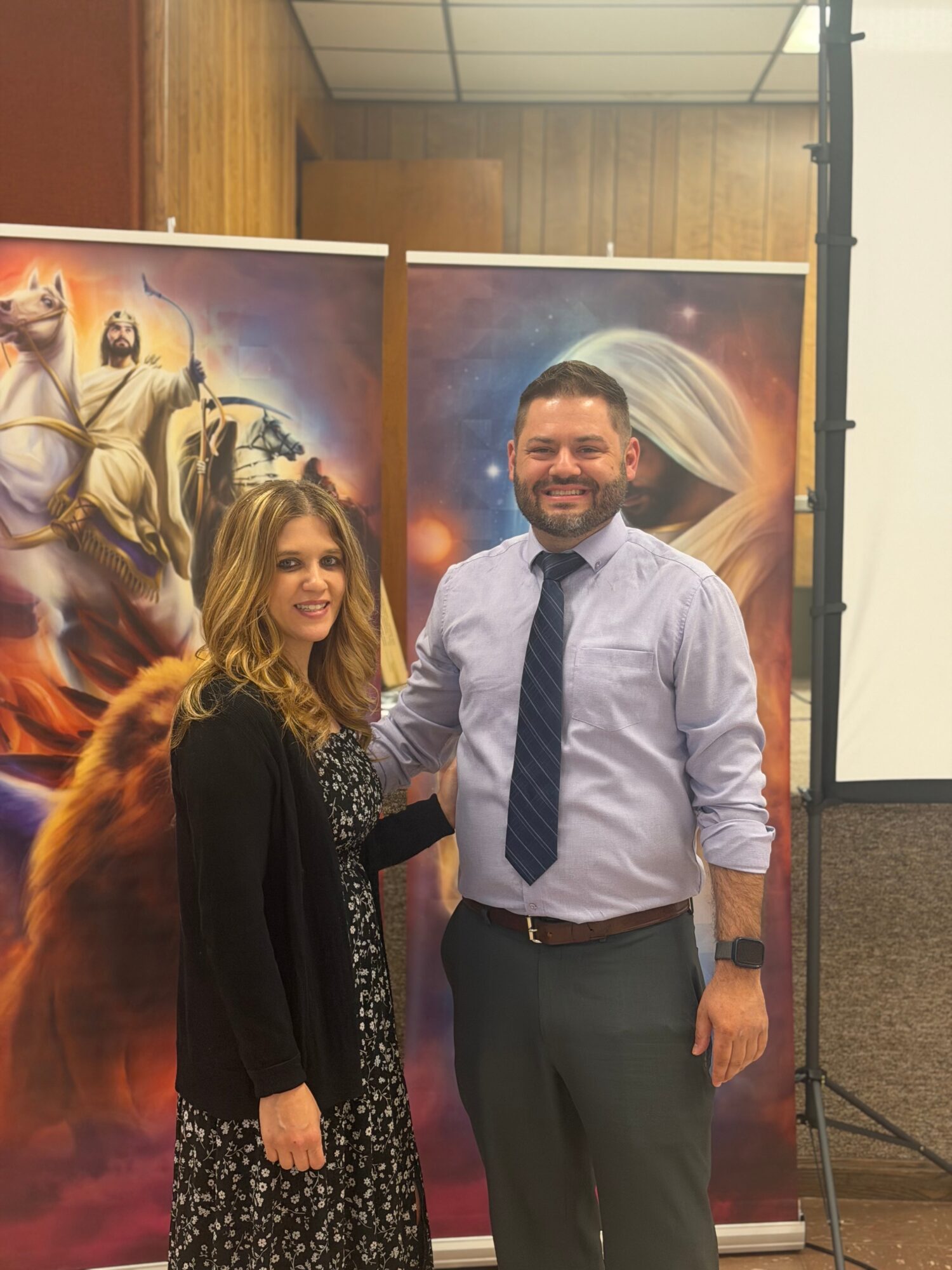 A man and woman stand together, smiling, in front of colorful fantasy-themed posters, indoors with wooden walls.