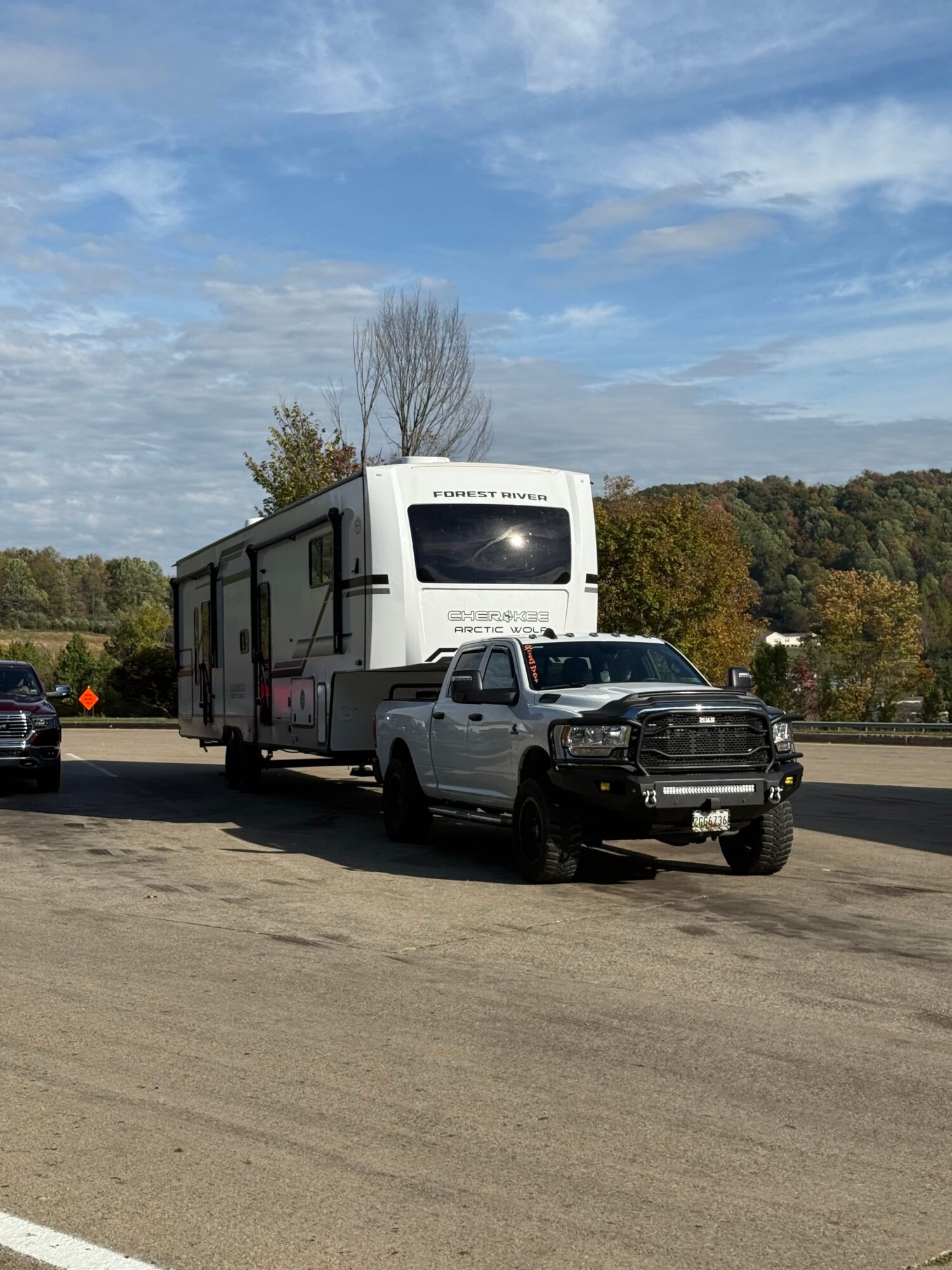 A white RV and a black pickup truck parked on a paved area with trees and a blue sky in the background.