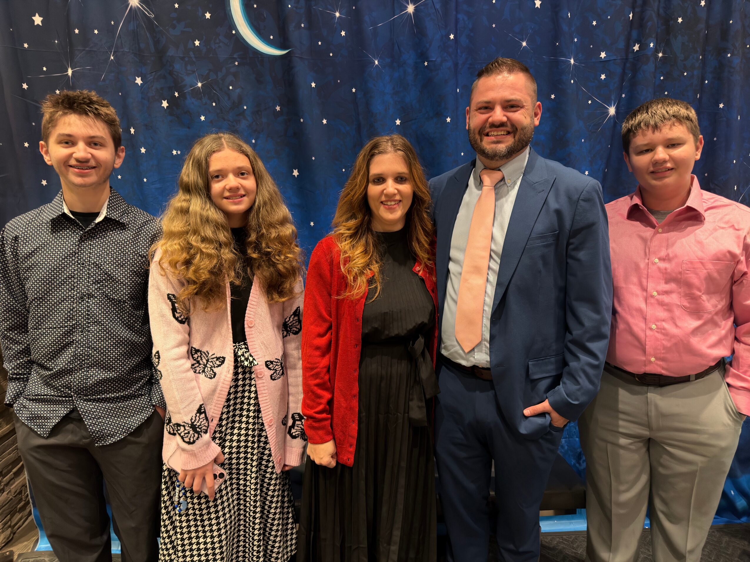 Group of five people standing in front of a starry night sky backdrop, smiling, with three young women and two men.