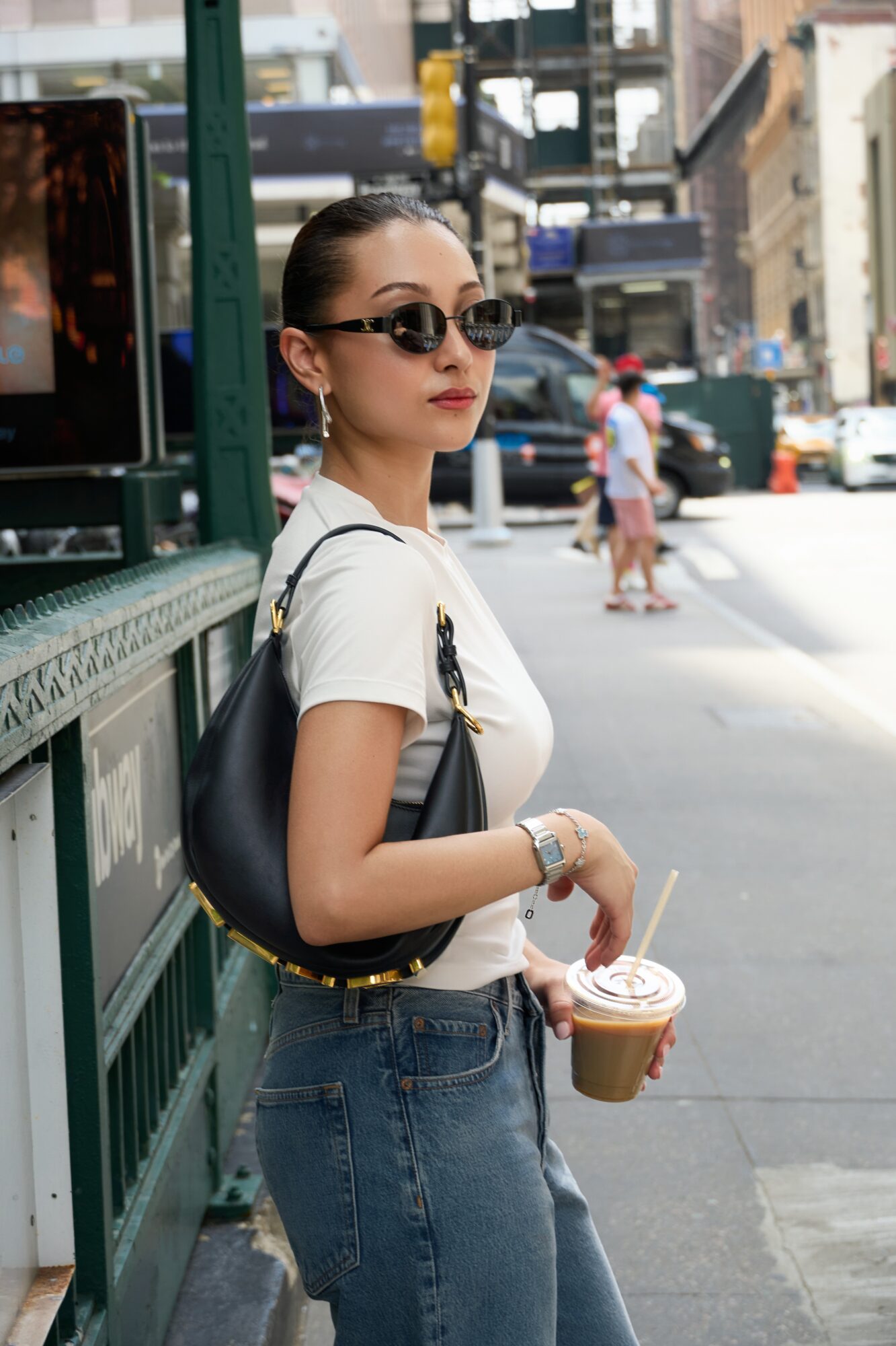 Young woman with sunglasses holding a coffee cup on city sidewalk, wearing a white t-shirt and jeans.